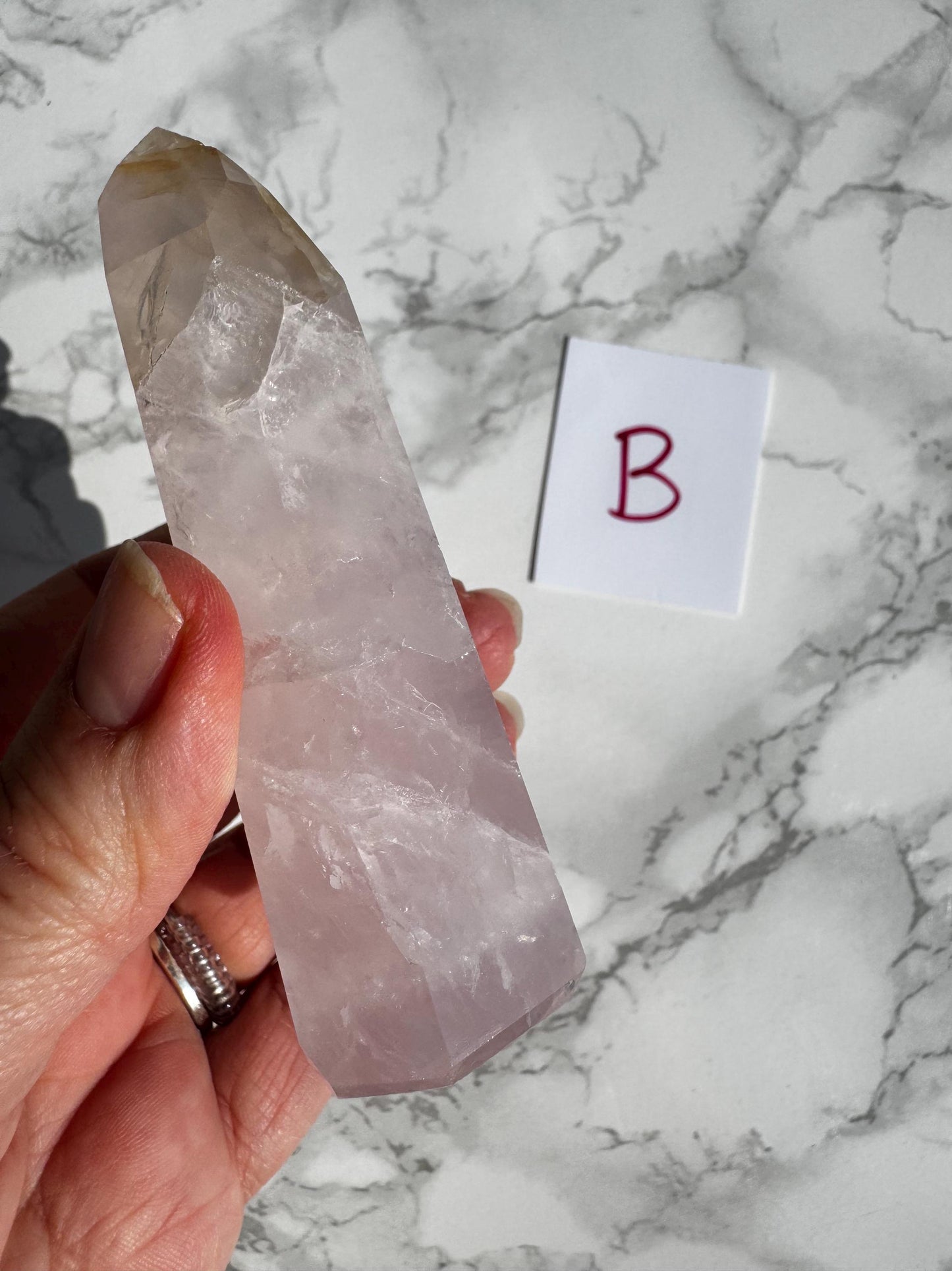 A hand holds a Nice Quality Rose Quartz Obelisk Tower from Brazil (3.6" - 3.8") beside a white card with "B" in red, all on a marble surface.