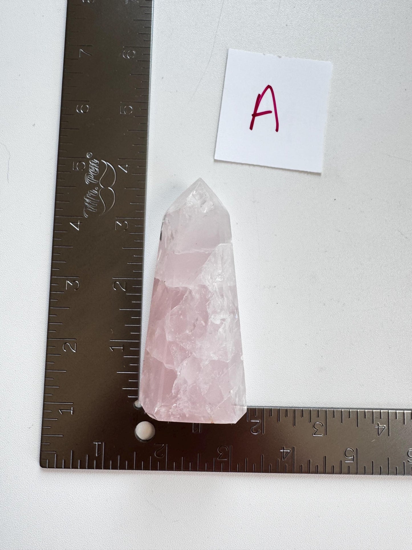 A Nice Quality Rose Quartz Obelisk Tower from Brazil (3.6"–3.8") rests between two L-shaped metal rulers, with a red "A" label nearby—an intriguing setup for healing crystal enthusiasts.