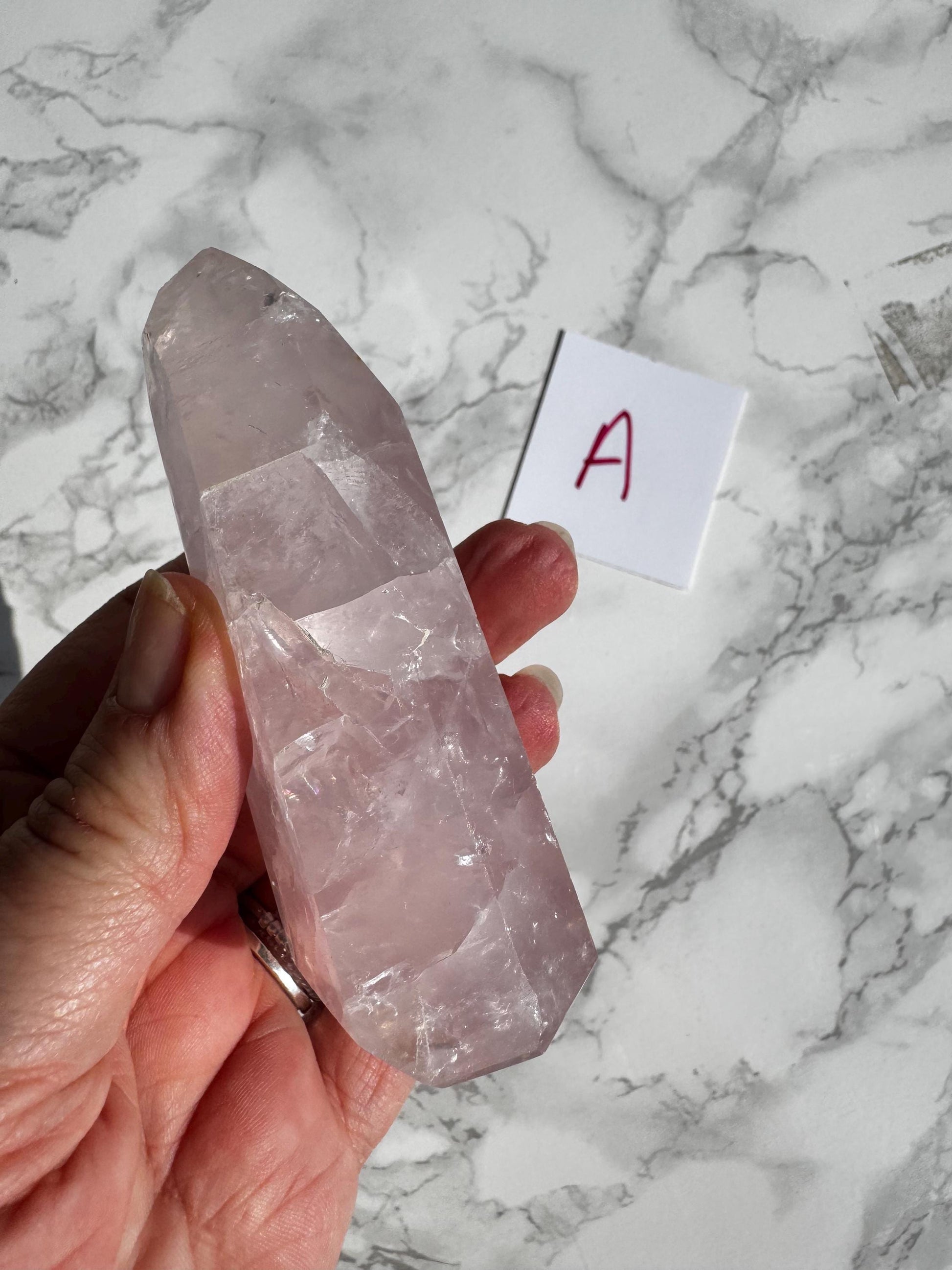 A hand holds a Nice Quality Rose Quartz Obelisk Tower from Brazil (3.6"–3.8") over marble, with a small white card labeled "A" nearby—ideal for lovers of healing crystals and elegant rose quartz towers.