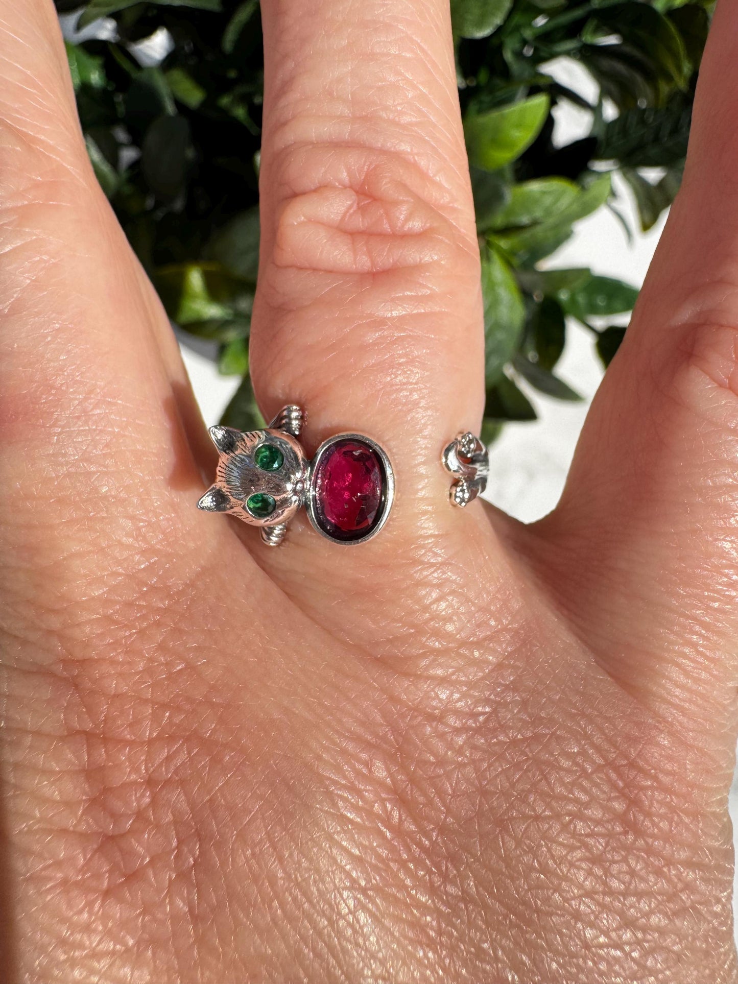 A Sterling Silver Cat Ring with a pink tourmaline gemstone (size 6.5, adjustable) features captivating feline details and sparkles on the finger; green leaves in the background highlight this distinctive crystal healing jewelry piece.