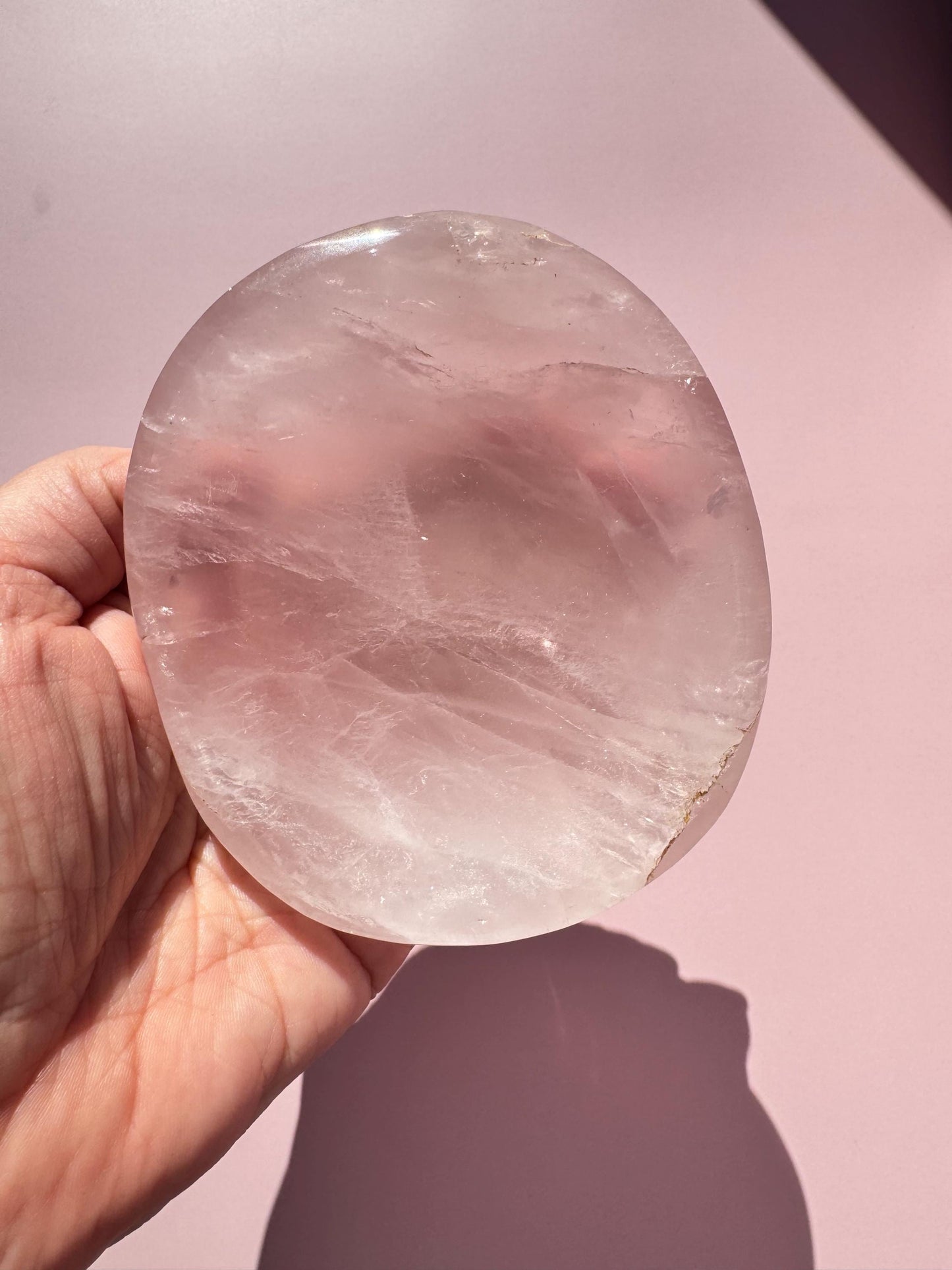 A hand holds the Rose Quartz Bowl - ITEM B, a large, flat pink crystal bowl from Madagascar, against a light pink background, highlighting its translucent beauty—perfect for holding small tumbles, jewelry, or trinkets.