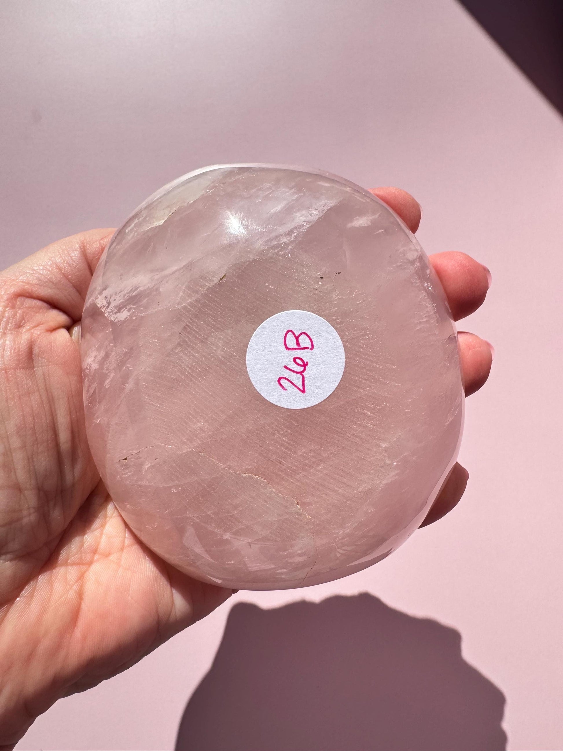 A hand holds the Rose Quartz Bowl - ITEM B, a polished pink crystal bowl perfect for small tumbles, jewelry, or trinkets. A white sticker reads "26B" in red ink as the bowl glows against a light pink background.