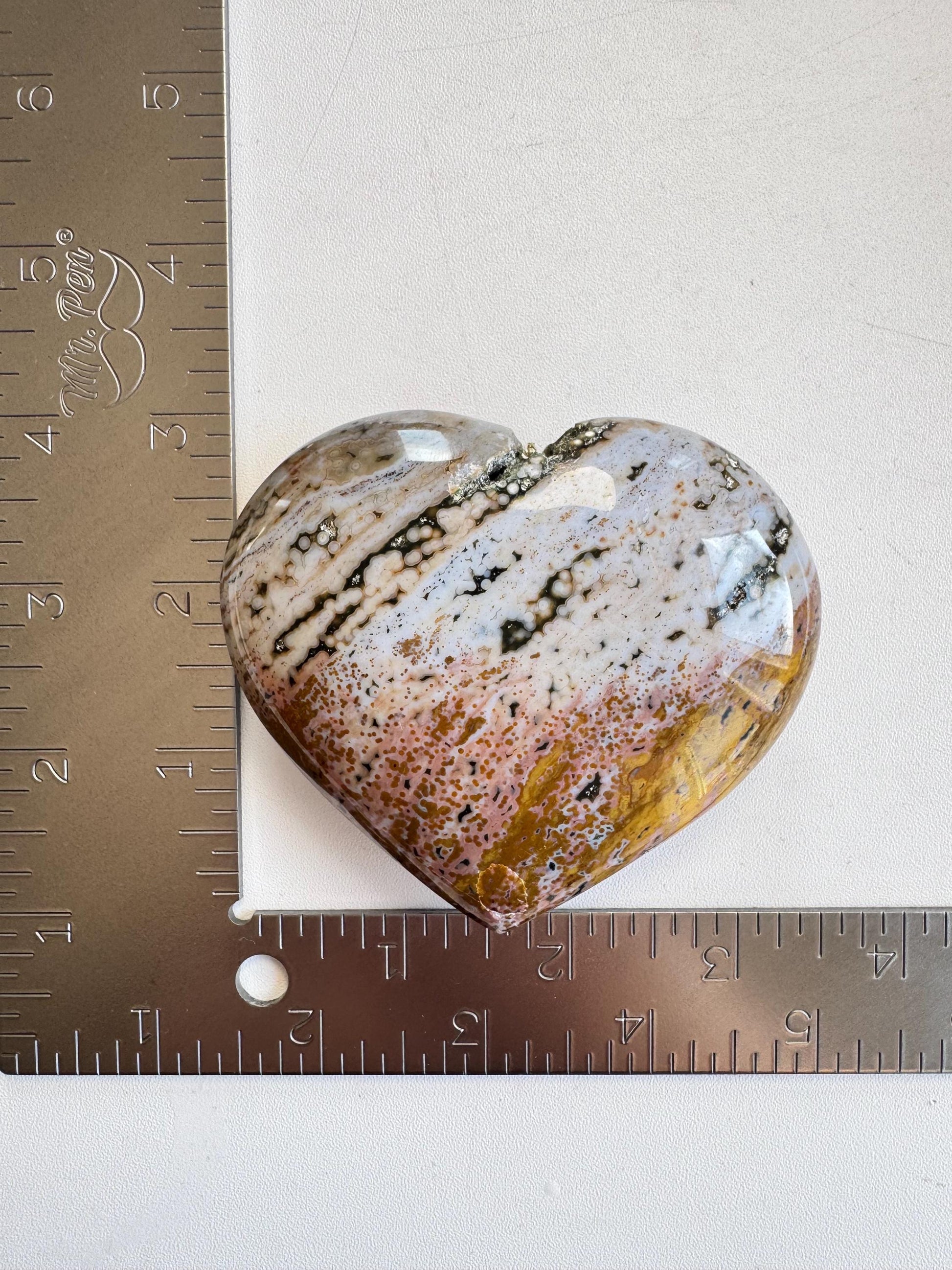The Ocean Jasper Heart from Madagascar - Item C, with brown, white, and gray patterns, is shown between two metal rulers forming a right angle to display its width of about 3 inches.