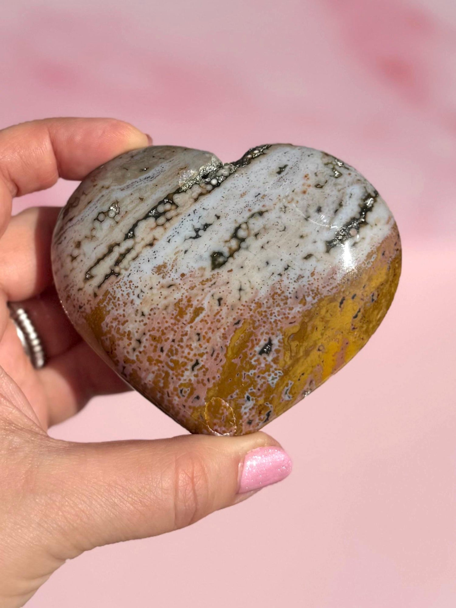 A hand holds the Ocean Jasper Heart from Madagascar - Item C, displaying white, brown, and yellow bands on a pink background. This crystal is valued for its healing properties.