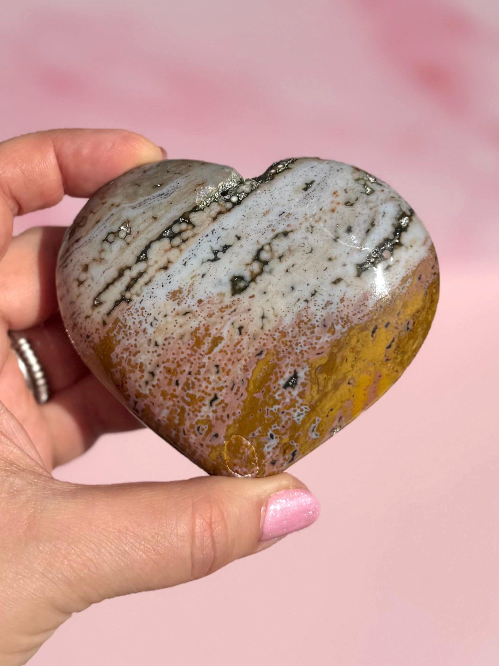 A hand holds the Ocean Jasper Heart from Madagascar - Item C, displaying white, brown, and yellow bands on a pink background. This crystal is valued for its healing properties.