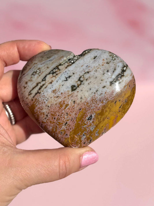 A hand holds the Ocean Jasper Heart from Madagascar - Item C, displaying white, brown, and yellow bands on a pink background. This crystal is valued for its healing properties.