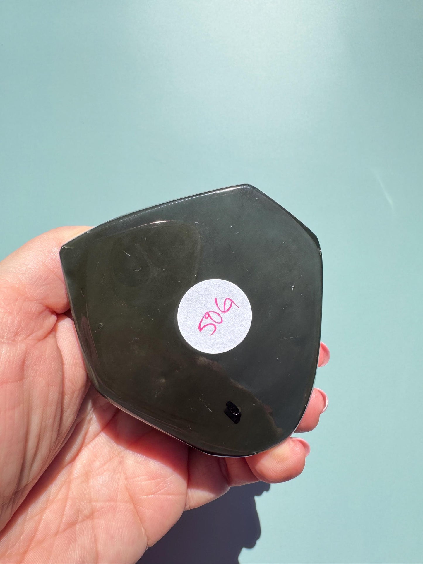 A hand holds the Rainbow Obsidian Heart Carving from Mexico (ITEM G), a dark, shield-shaped stone with a white sticker labeled “506” in pink ink, set against a light blue background.