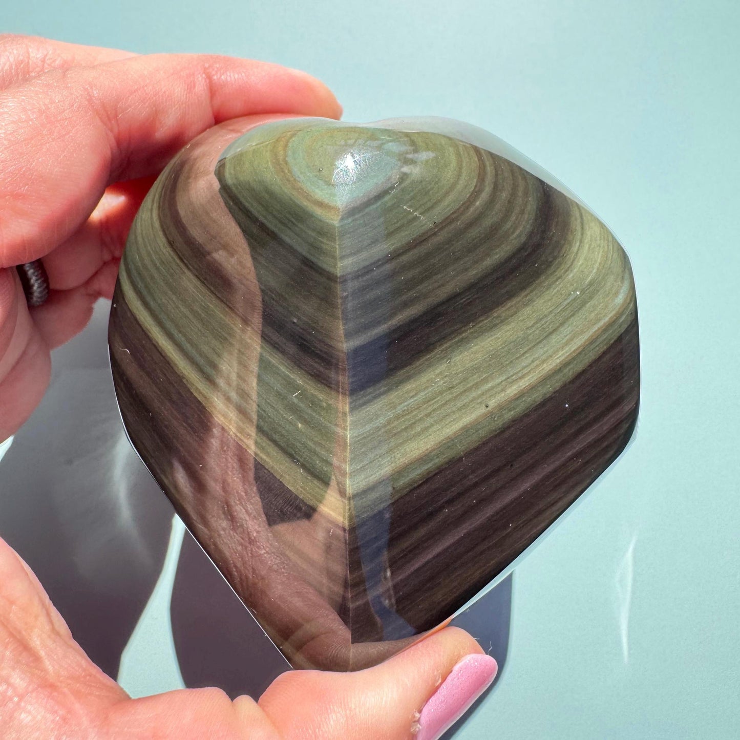 A hand holds the Rainbow Obsidian Heart Carving from Mexico (ITEM G), a polished protective stone with layered green and brown bands—an elegant piece for any healing crystals collection—set against a light green background.