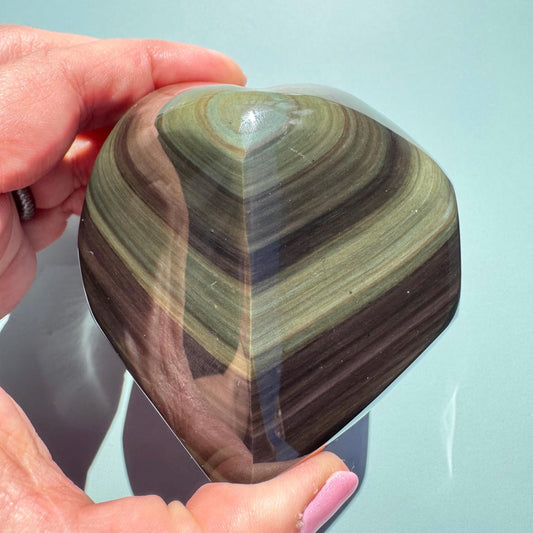 A hand holds the Rainbow Obsidian Heart Carving from Mexico (ITEM G), a polished protective stone with layered green and brown bands—an elegant piece for any healing crystals collection—set against a light green background.
