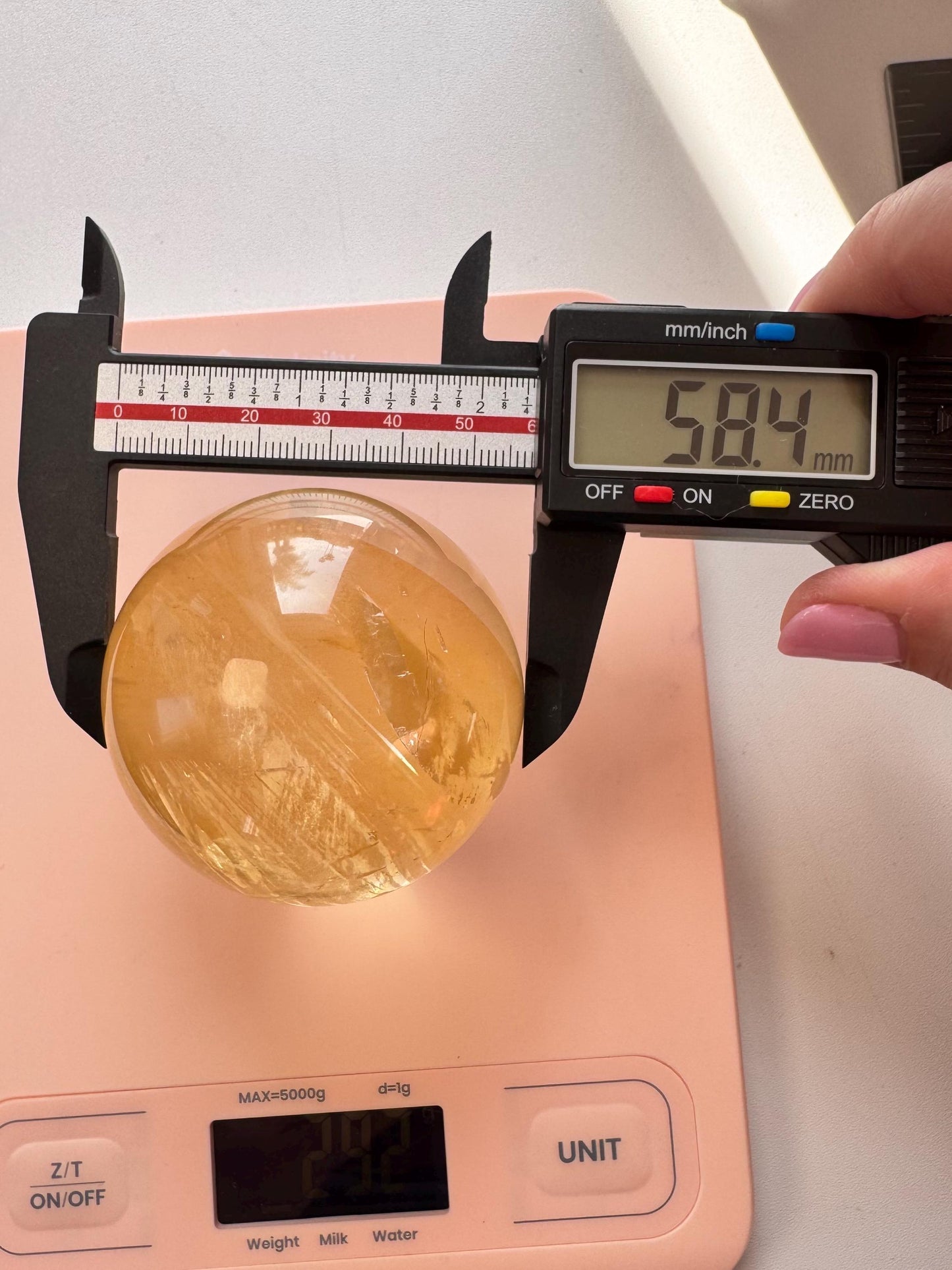 A Honey Calcite Sphere (58 mm diameter) appears yellow and measures 58.4 mm with a digital caliper when placed on a pink digital scale.