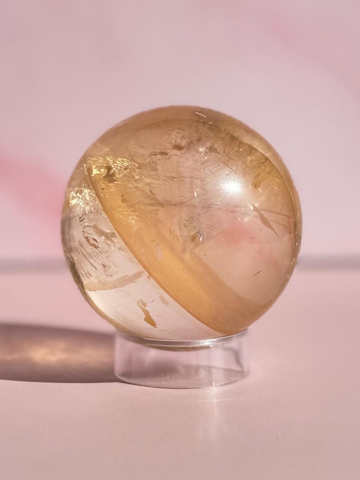 The 58 mm Honey Calcite Sphere features golden clarity and internal striations, displayed on a transparent stand with a pink background—an inspiring crystal for motivation and clarity.