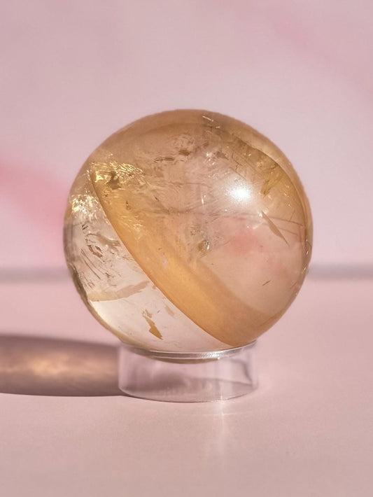 The 58 mm Honey Calcite Sphere features golden clarity and internal striations, displayed on a transparent stand with a pink background—an inspiring crystal for motivation and clarity.