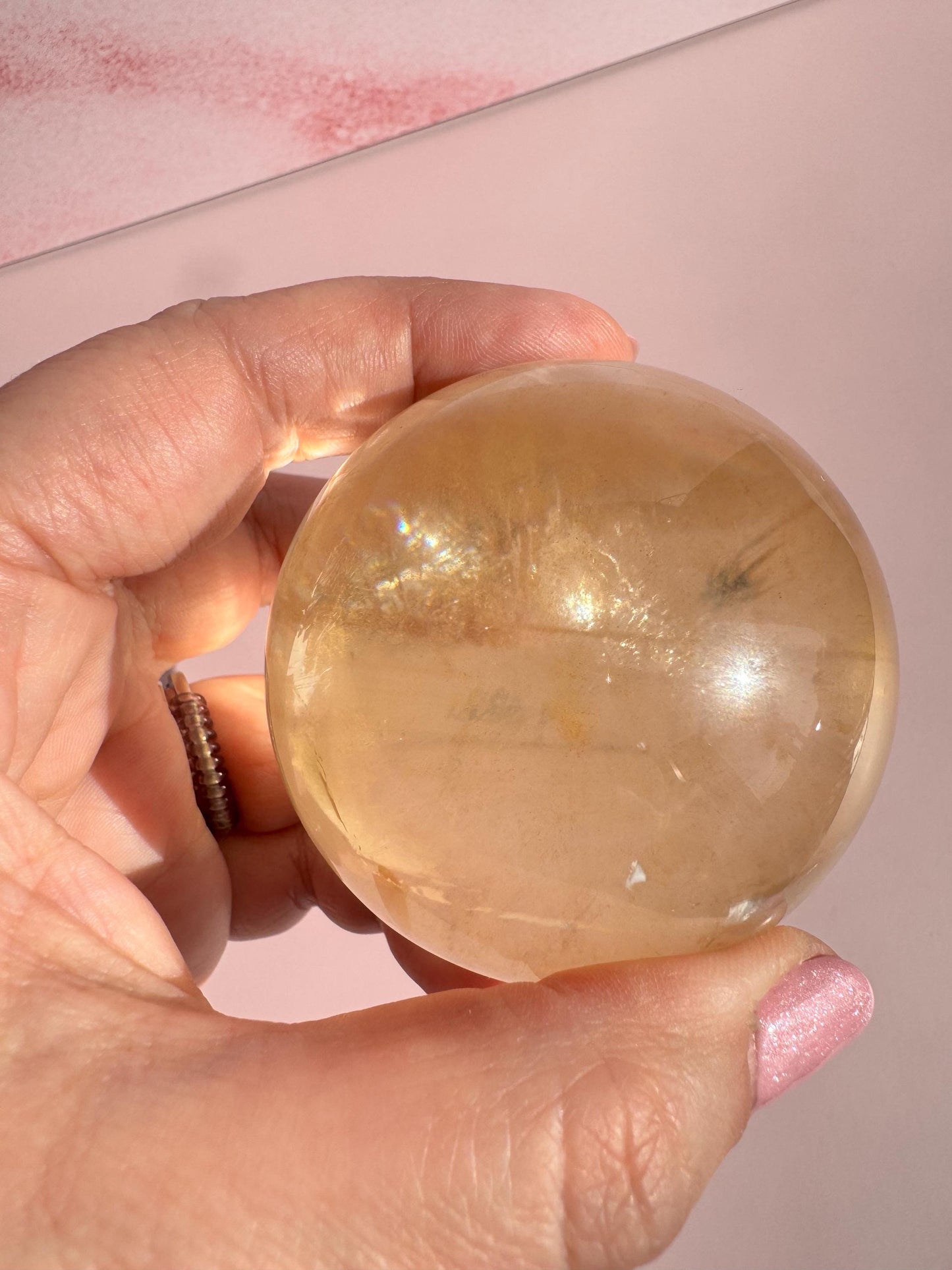 A hand holds the 58 mm Honey Calcite Sphere—a smooth, round crystal known for boosting motivation—against a soft pink background.