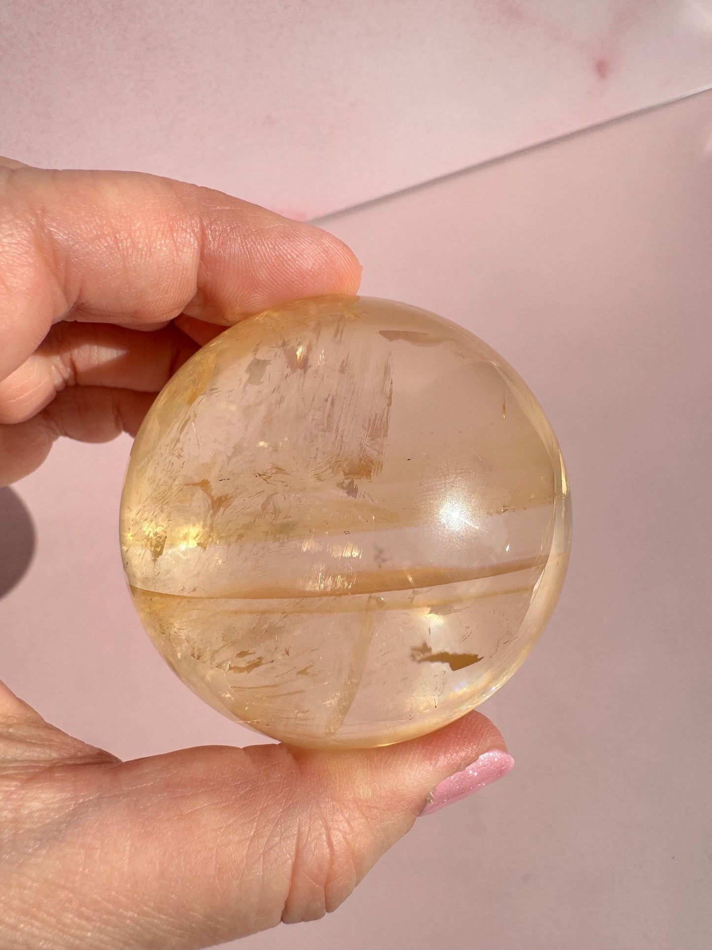 A hand holds the Honey Calcite Sphere (58 mm diameter), a crystal known for motivation and positive energy, against a pale pink background.