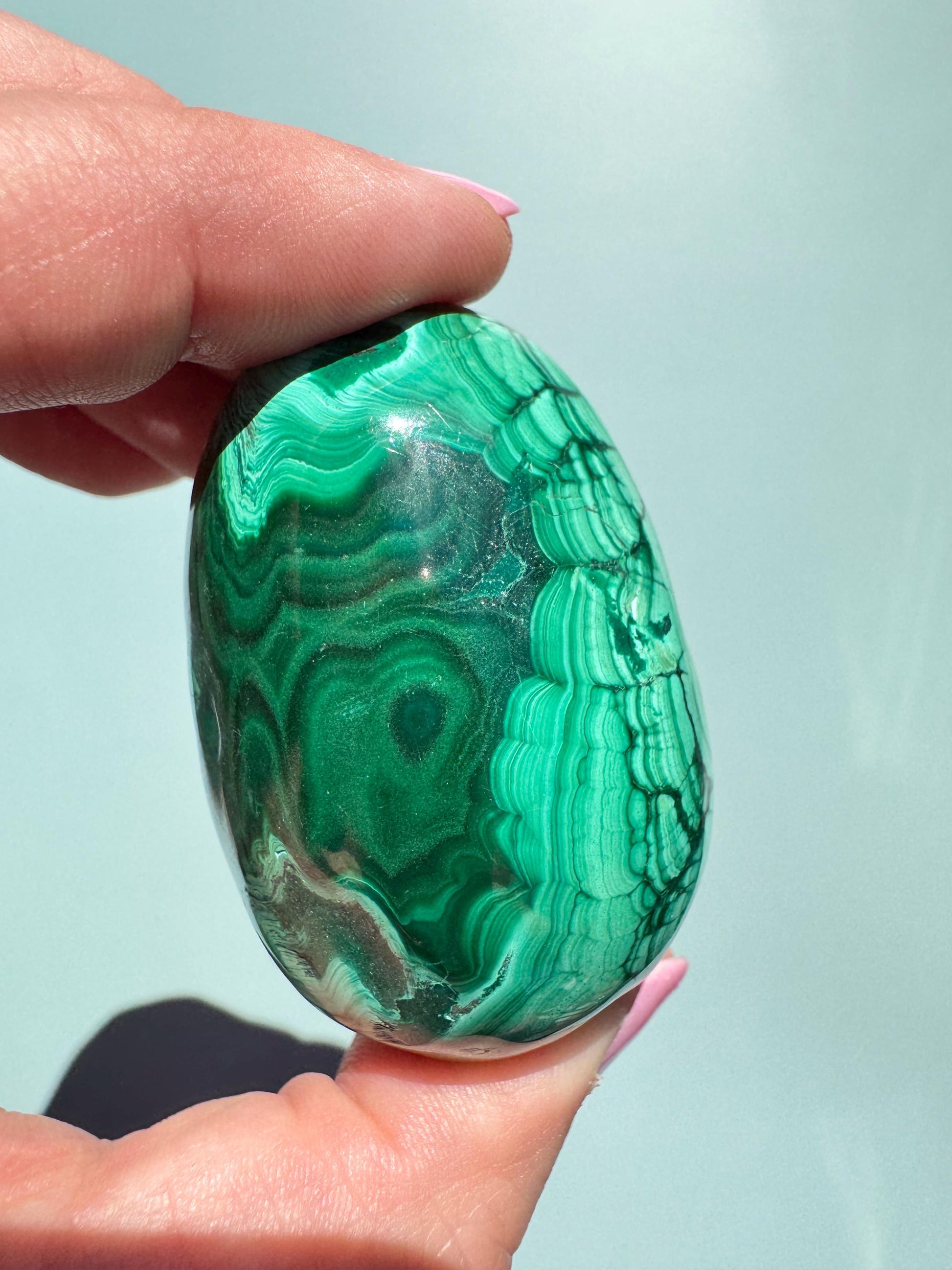 A hand holds the Malachite Egg-Shaped Carving (ITEM C), a polished green stone with distinctive banded patterns, displayed against a light background.