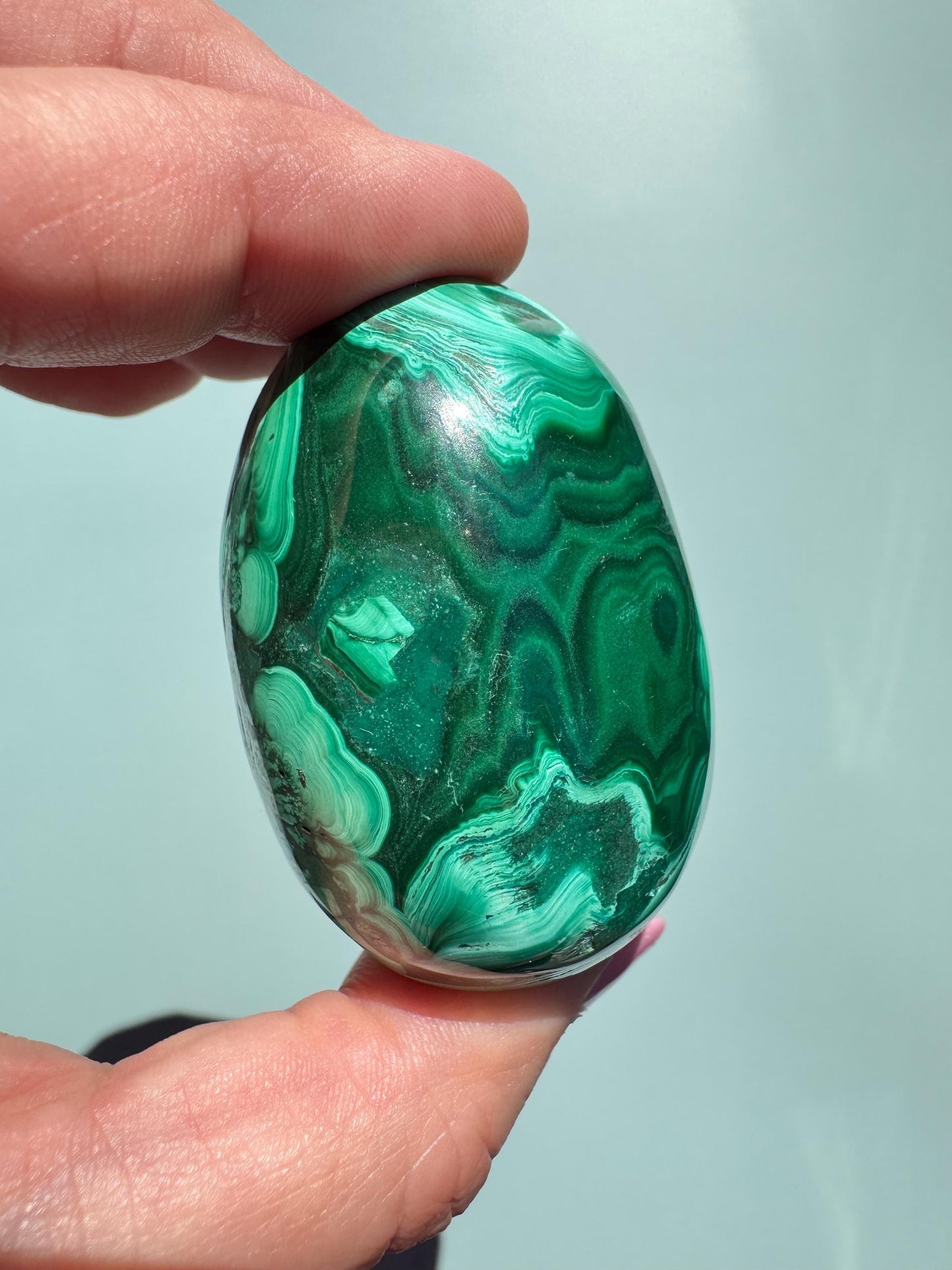 A hand holds the Malachite Egg-Shaped Carving (ITEM C), prized among healing crystals for its polished green surface and swirling, layered patterns, against a plain light background.