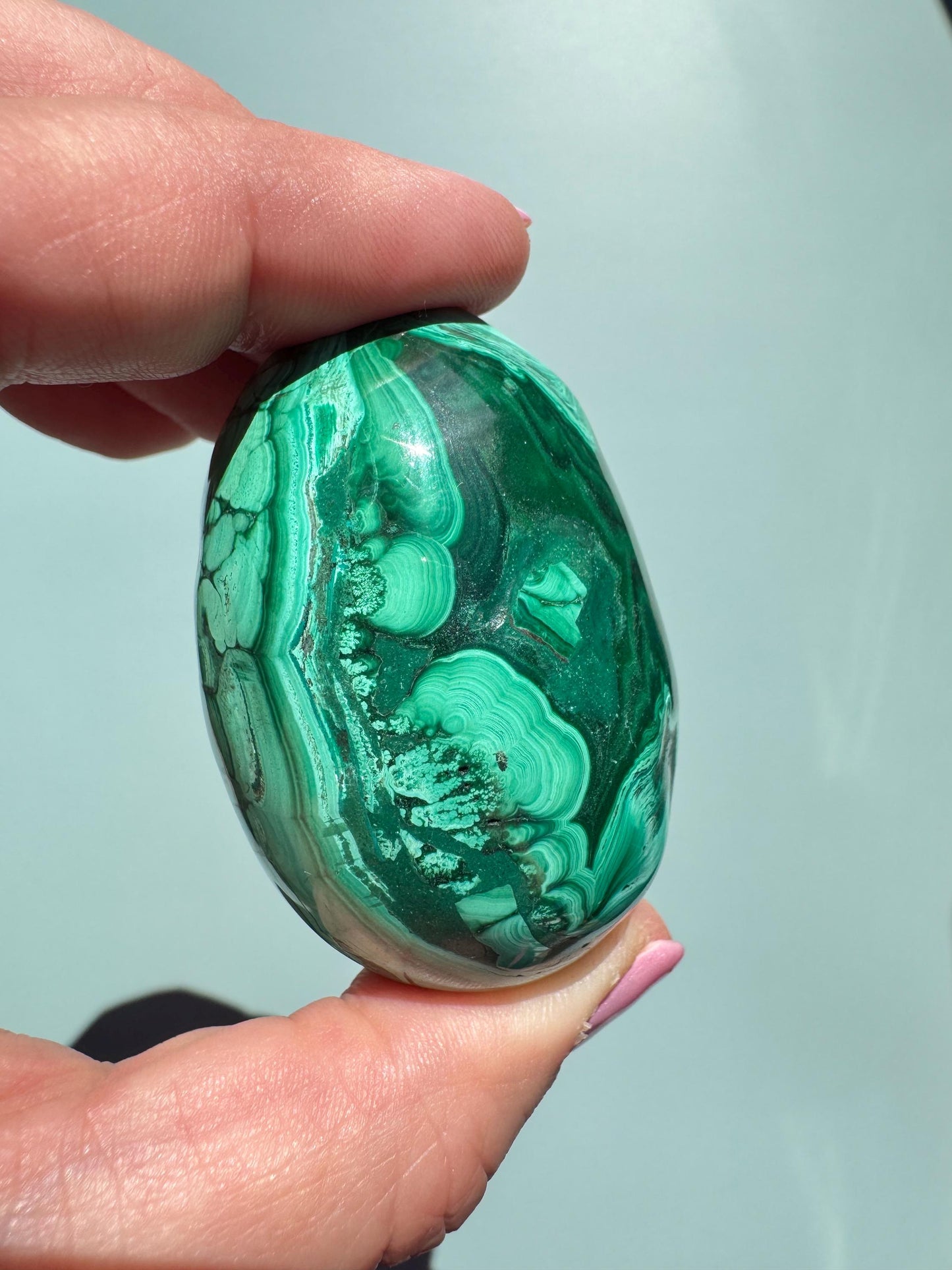 A hand holds the Malachite Egg-Shaped Carving (ITEM C), showcasing its polished green surface with unique light and dark bands against a plain light background.