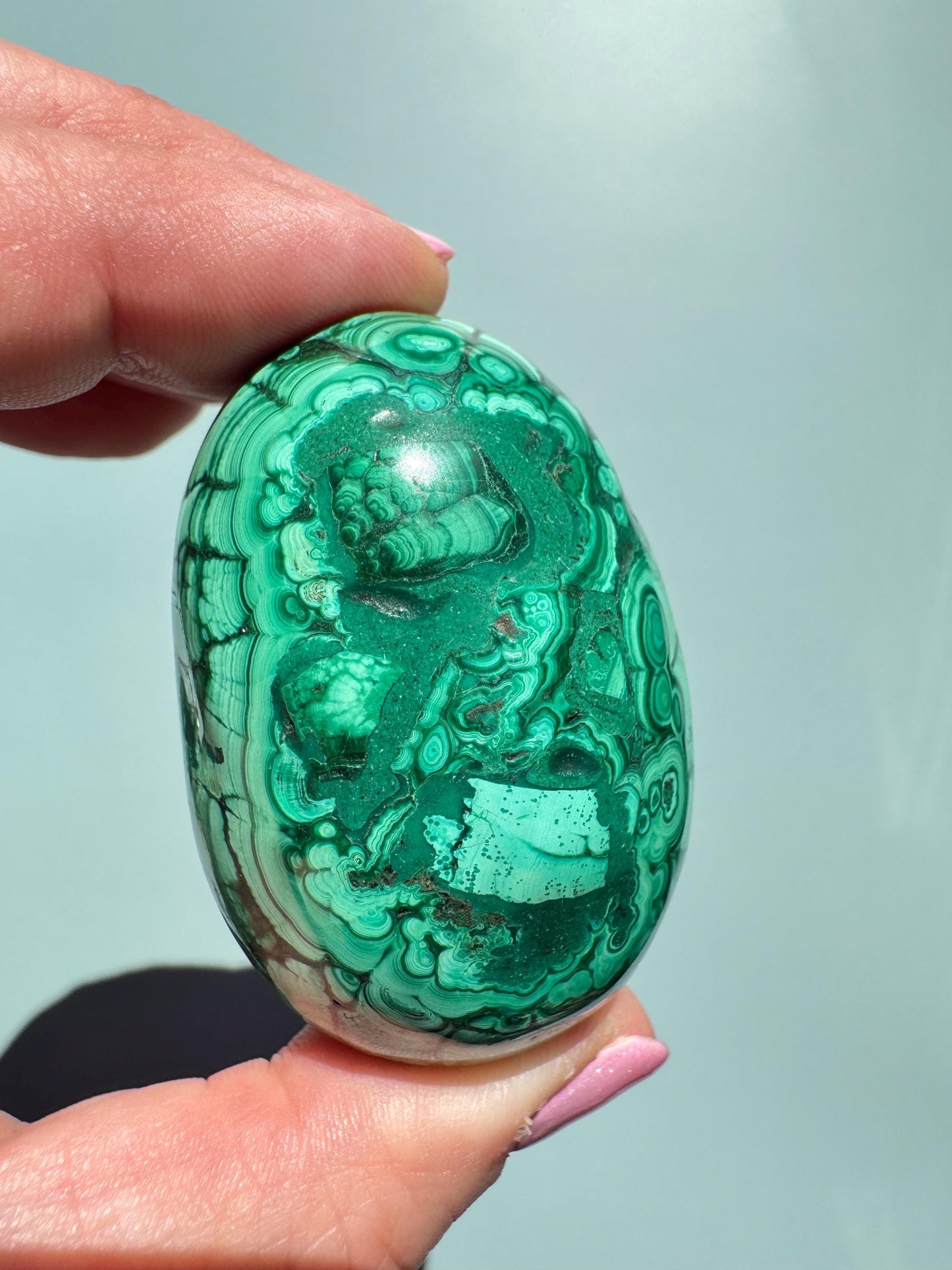 A hand holds the Malachite Egg-Shaped Carving (ITEM C), a polished green crystal renowned for transformation, its swirling banded patterns striking against a plain light background.