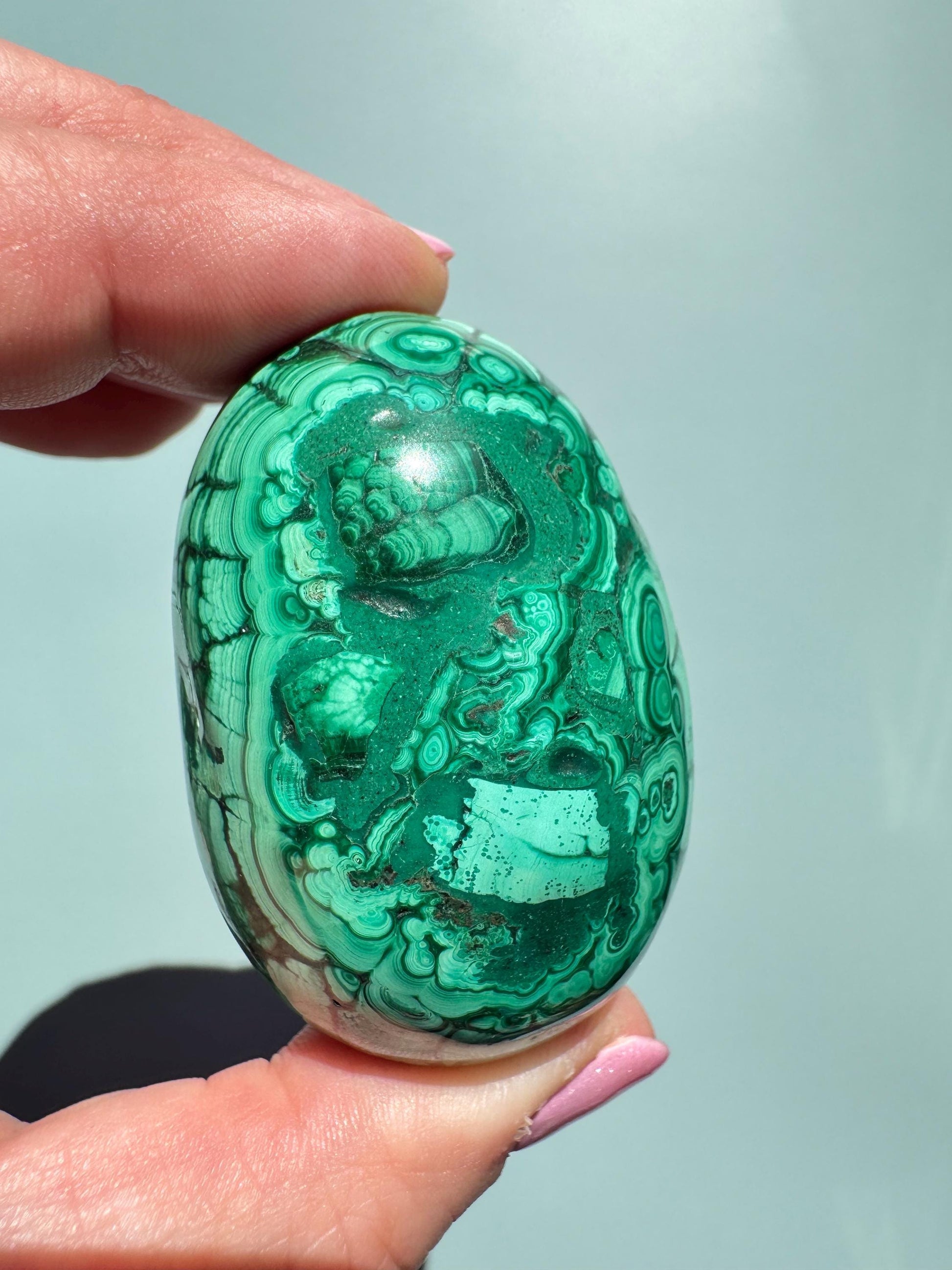 A hand holds the Malachite Egg-Shaped Carving (ITEM C), a polished green crystal renowned for transformation, its swirling banded patterns striking against a plain light background.