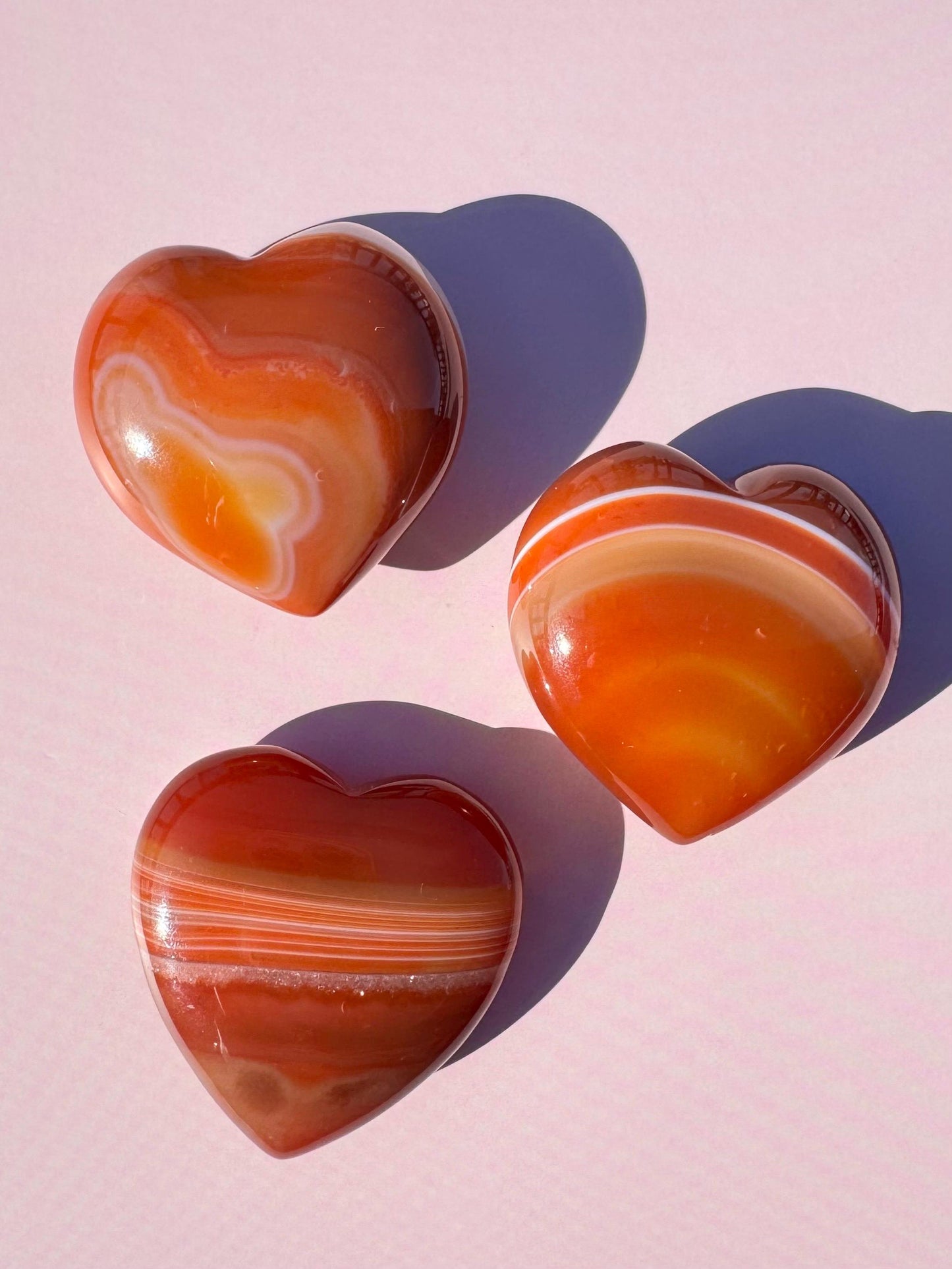 Three polished Small Carnelian Heart Carvings (YOU CHOOSE) are arranged on a pastel pink surface, casting soft shadows and evoking the look of healing crystals.