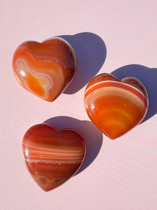 Three polished Small Carnelian Heart Carvings (YOU CHOOSE) are arranged on a pastel pink surface, casting soft shadows and evoking the look of healing crystals.