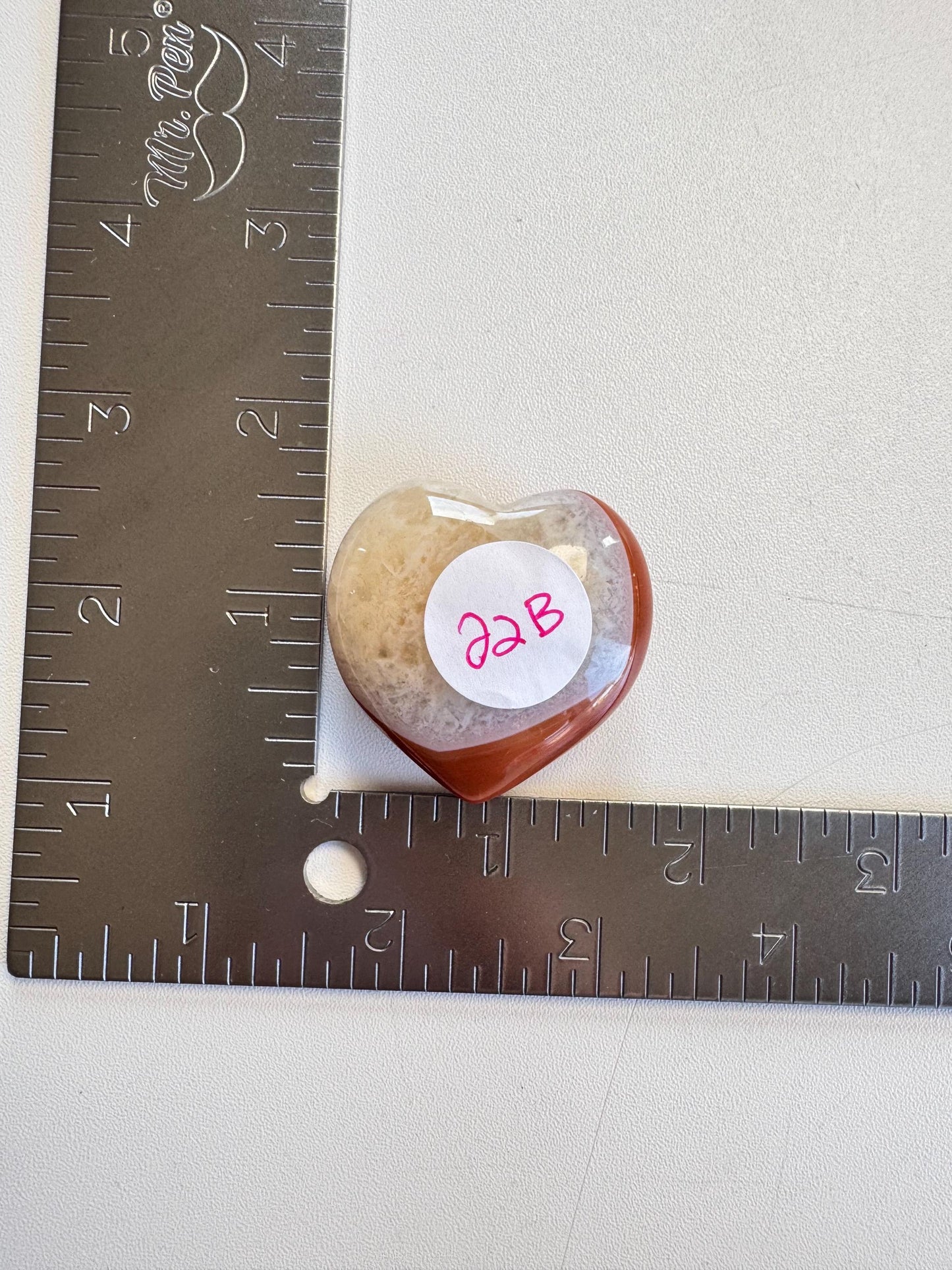 A Small Carnelian Heart Carving - YOU CHOOSE sits at the intersection of two metal rulers on a white surface, its vivid energy and charm beautifully displayed.