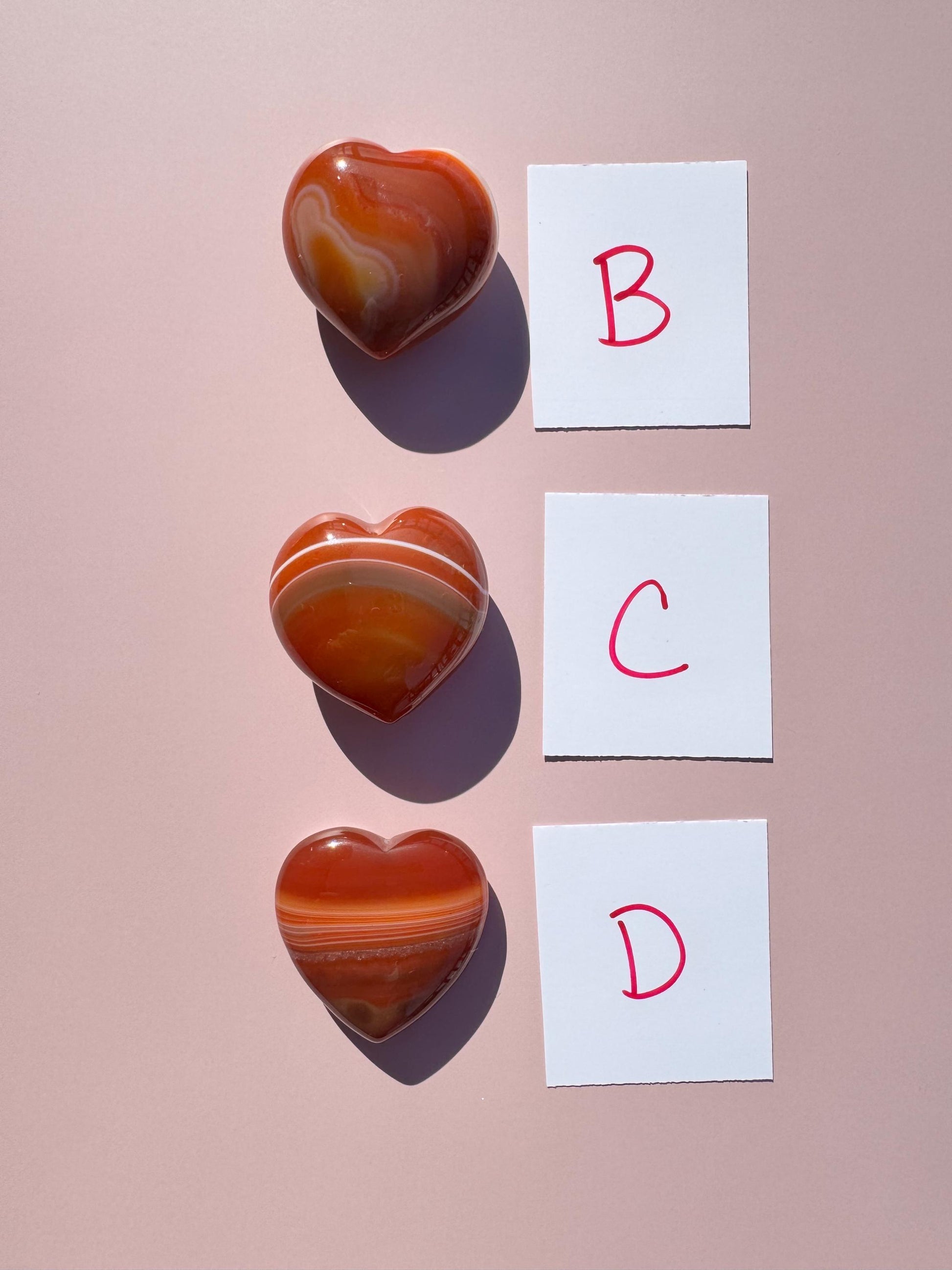 A Small Carnelian Heart Carving - YOU CHOOSE is displayed beside cards labeled B, C, and D on a pink surface, radiating the energizing stone’s warm vibes.