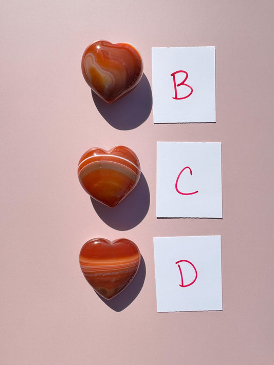 A Small Carnelian Heart Carving - YOU CHOOSE is displayed beside cards labeled B, C, and D on a pink surface, radiating the energizing stone’s warm vibes.