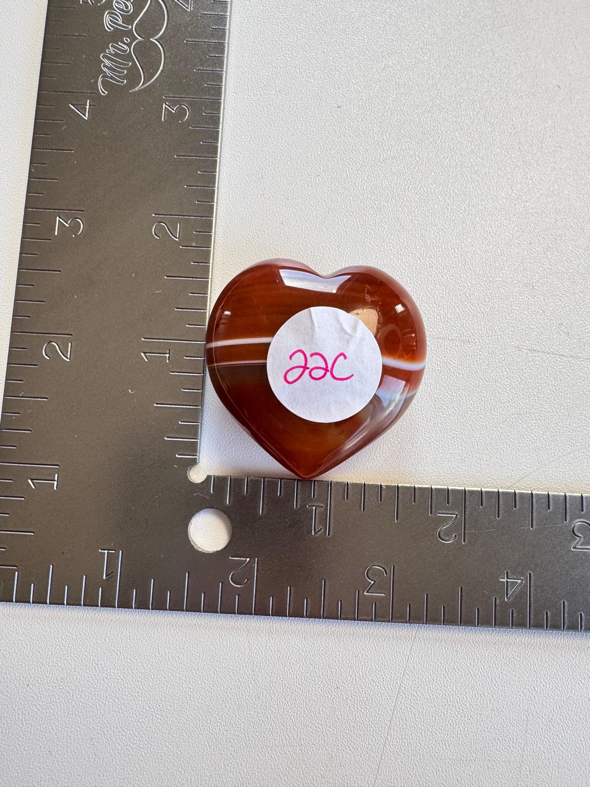 A Small Carnelian Heart Carving - YOU CHOOSE, with a "22C" sticker, sits at the intersection of two inch-marked metal rulers on a white surface—an energizing crystal popular among healing stones.