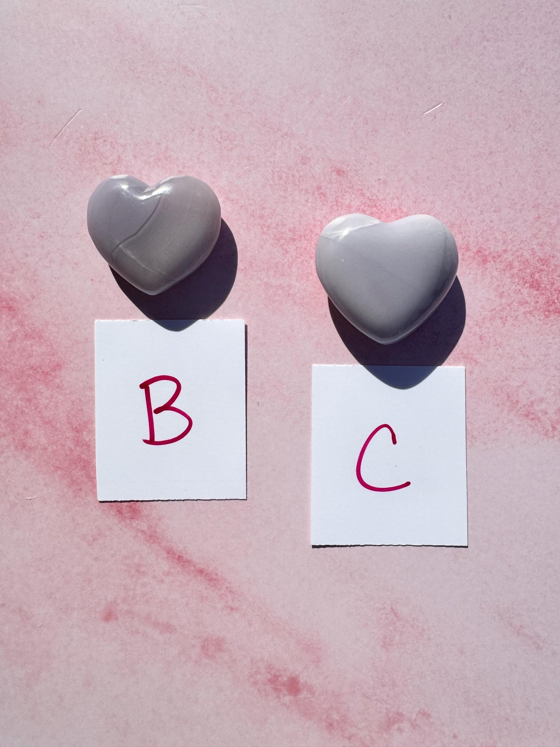 Two Lavender Fluorite Heart Carvings from Mexico, labeled "B" and "C," rest on a pink surface, each above a white card with a red letter.