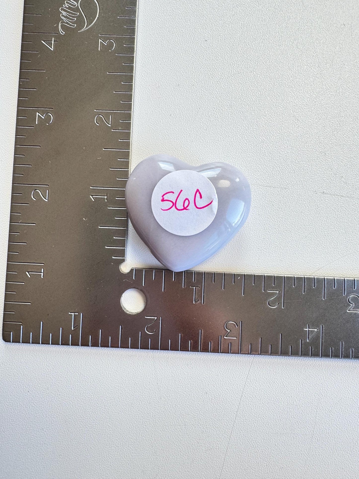 A Lavender Fluorite Heart Carving from Mexico (YOU CHOOSE), with a "56C" sticker, sits on a metal ruler marked in inches on a white surface.