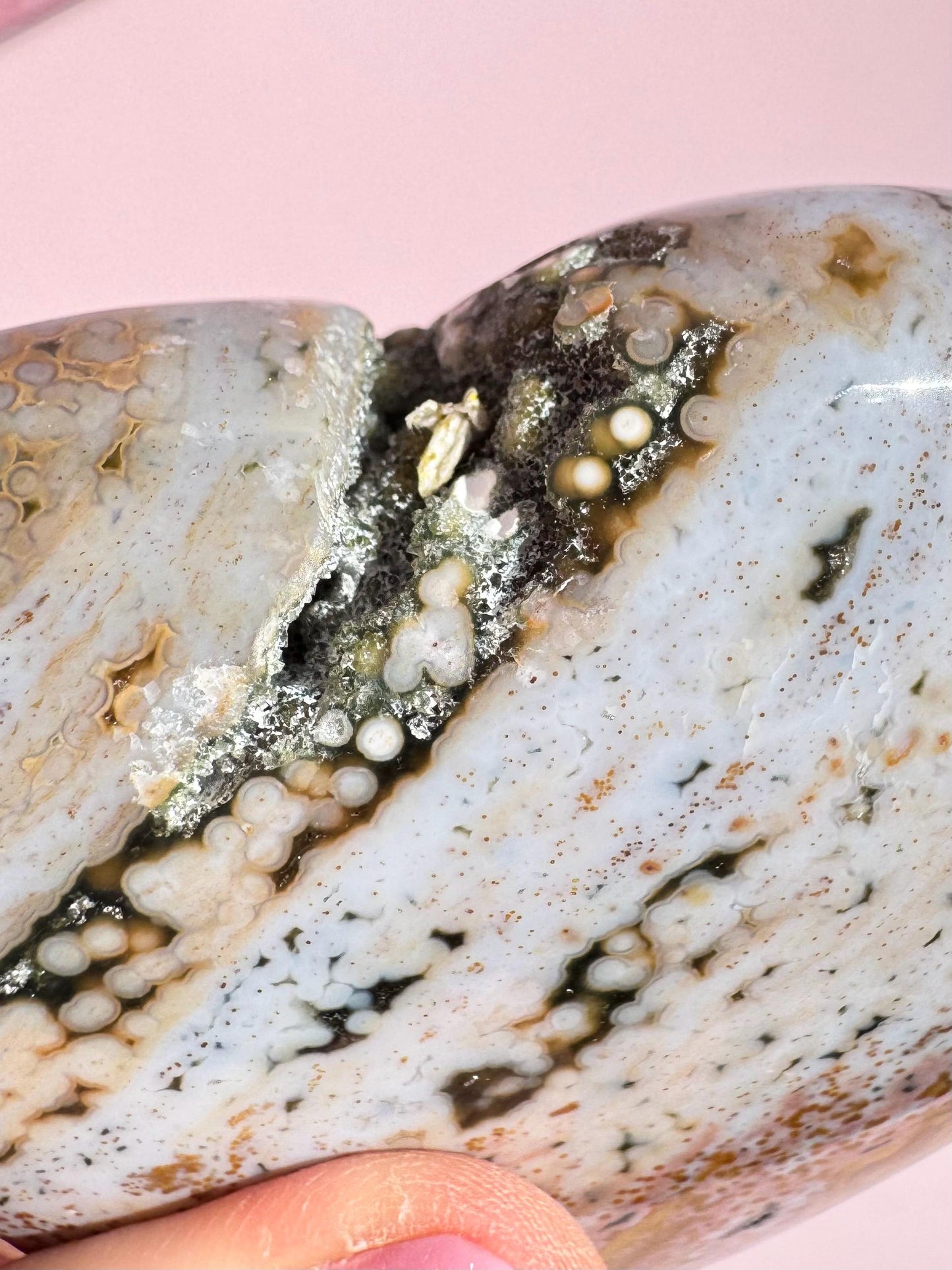 A close-up of the Ocean Jasper Heart from Madagascar - Item C, featuring layered patterns and small circular mineral formations, is delicately held between fingers against a pink background.