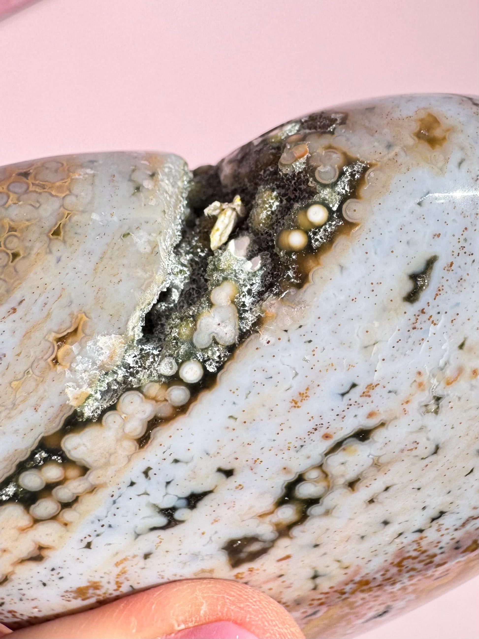 A close-up of the Ocean Jasper Heart from Madagascar - Item C, featuring layered patterns and small circular mineral formations, is delicately held between fingers against a pink background.