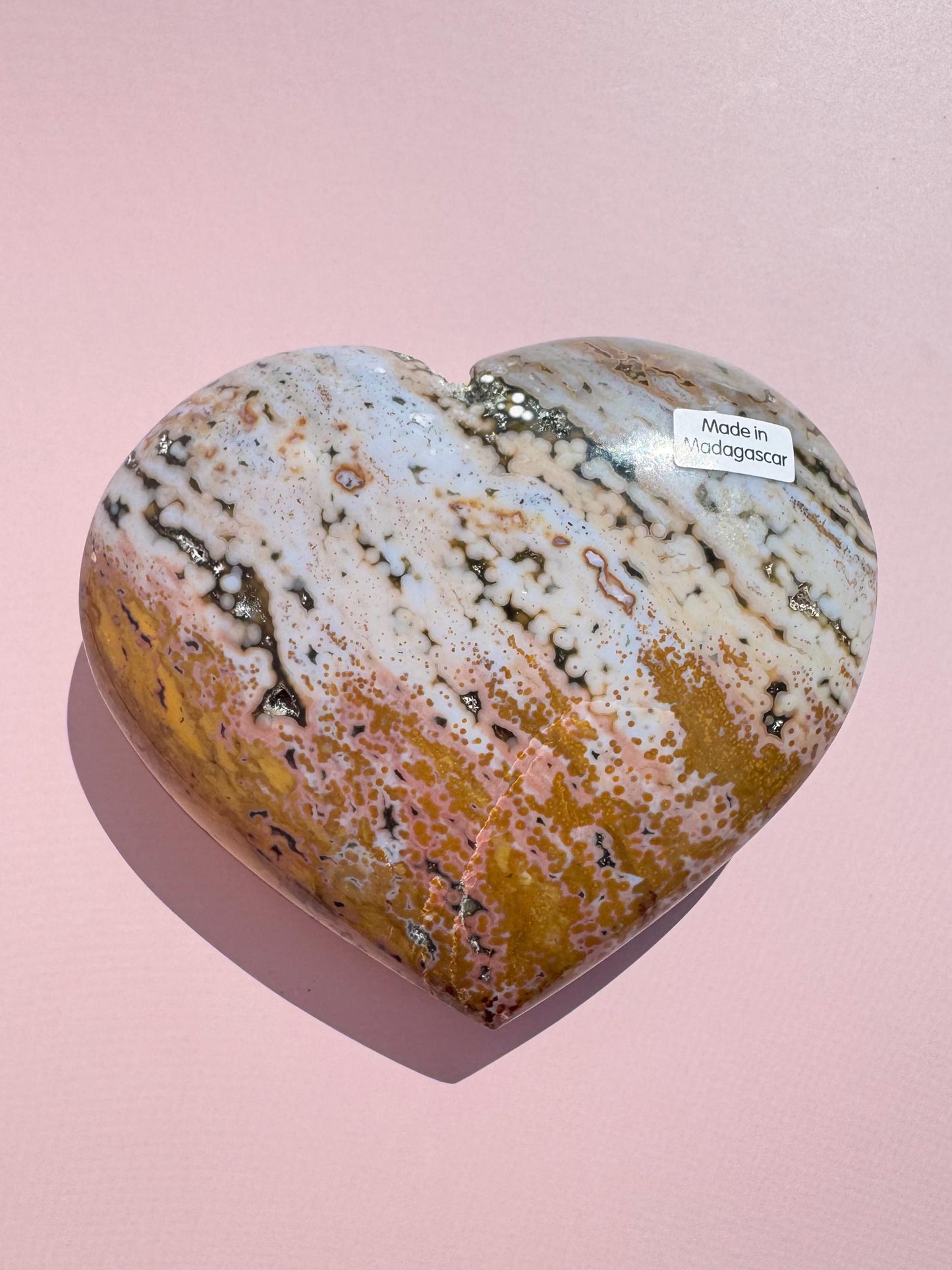 An Ocean Jasper Heart from Madagascar - Item C, featuring white, beige, and brown patterns, rests on a pink surface. A small "Made in Madagascar" sticker is attached.