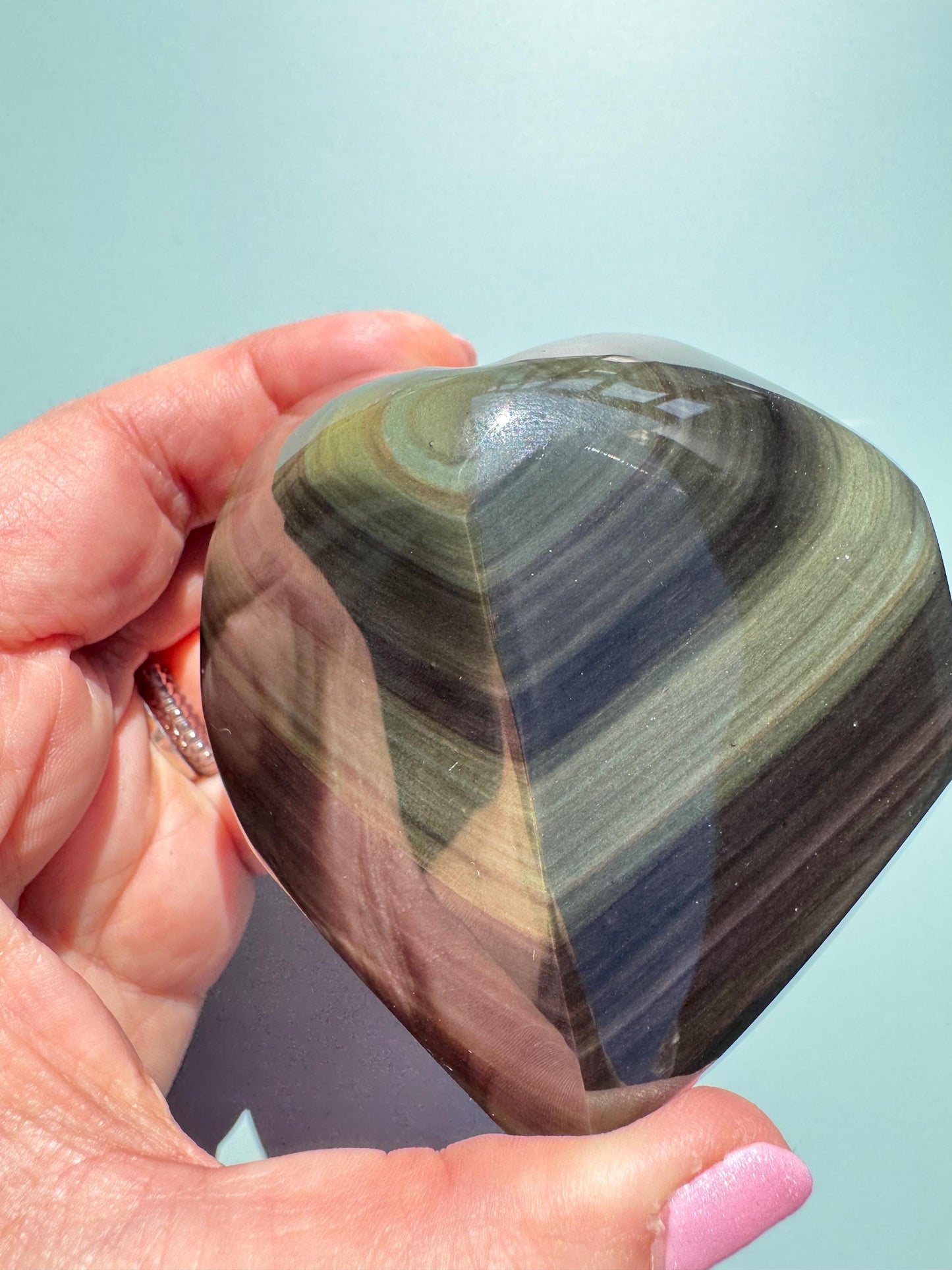 A hand holds a polished Rainbow Obsidian Heart Carving from Mexico (ITEM G), displaying layered green and brown bands like healing crystals, set against a light blue background.