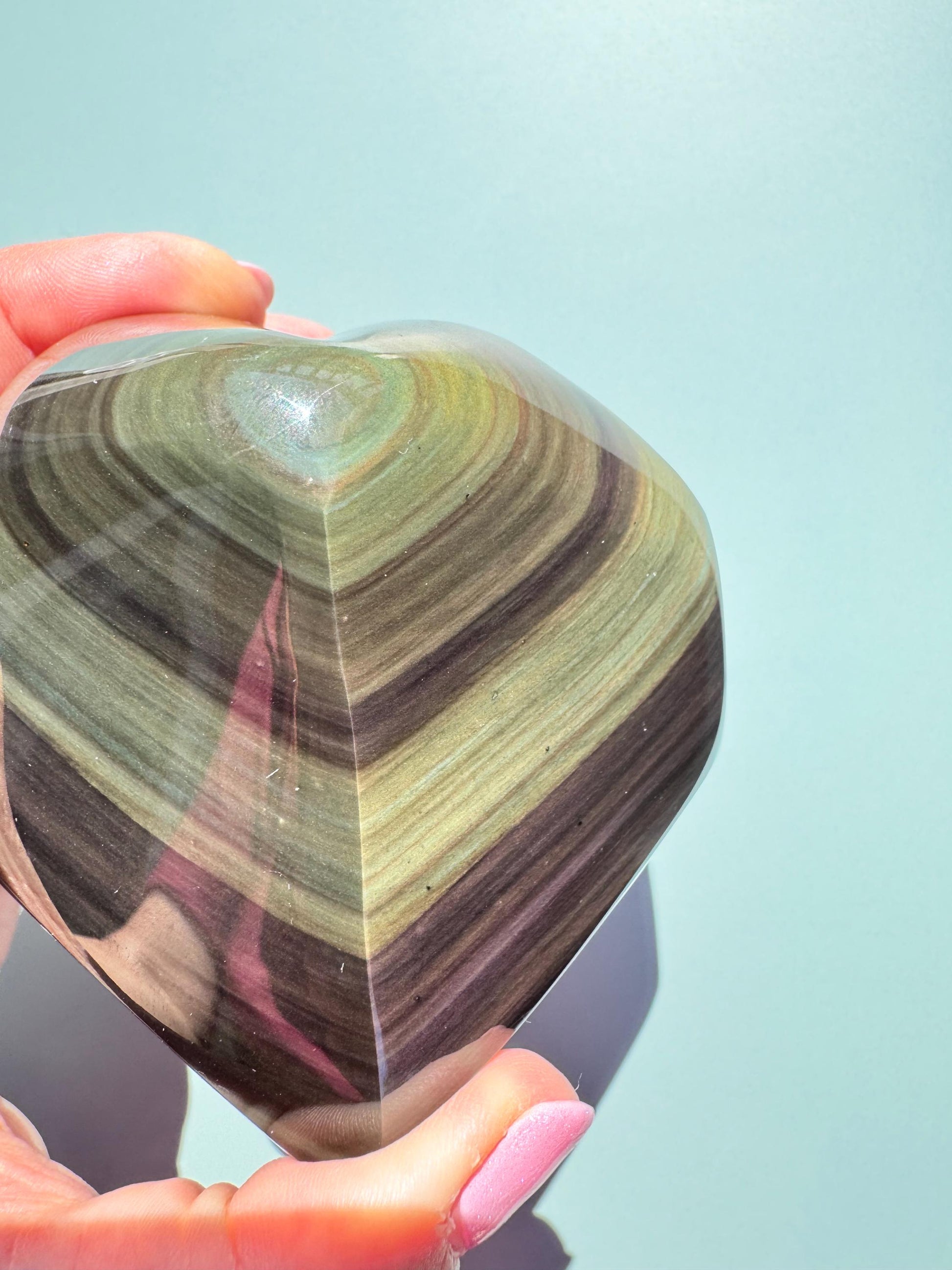 A hand holds a polished Rainbow Obsidian Heart Carving from Mexico (ITEM G), showing its natural green and brown banded layers against a light blue background.