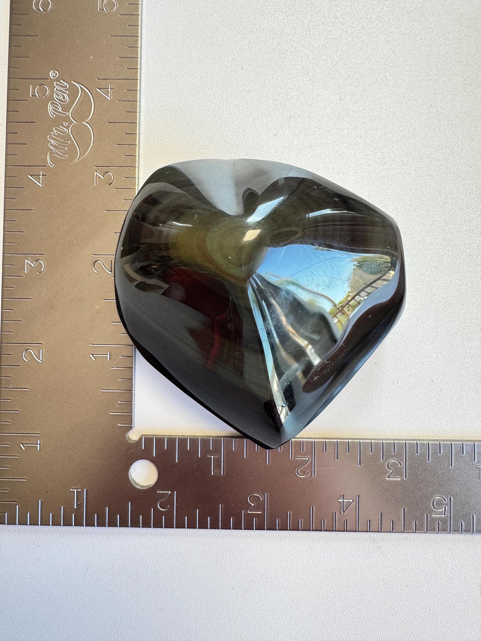 A polished Rainbow Obsidian Heart Carving from Mexico (ITEM G) sits on a white surface at the intersection of two metal rulers forming an L shape, measuring about 2.5 inches wide.
