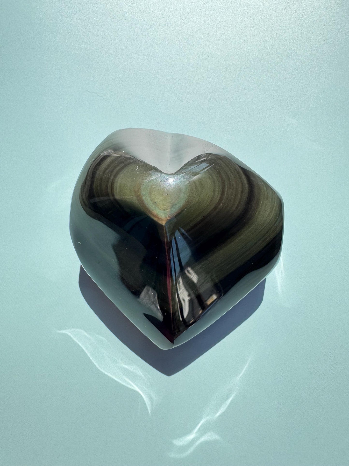 The Rainbow Obsidian Heart Carving - From Mexico (ITEM G) features dark, reflective bands and a smooth surface, highlighting its protective qualities as it rests on a light blue background.