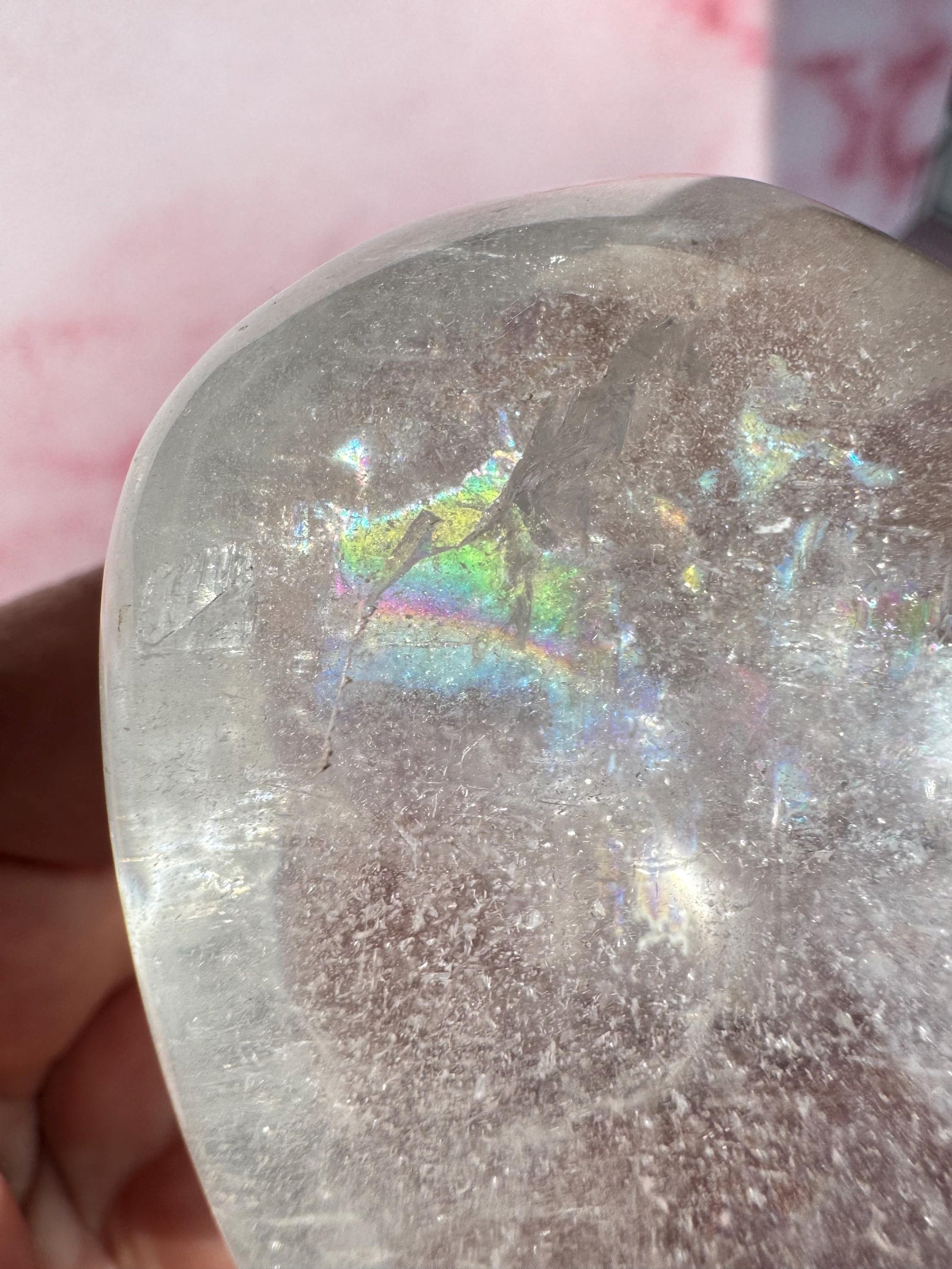 Close-up of our Clear Quartz Heart Carving – Very High Clarity Crystal, showing internal fractures and rainbow reflections, held in hand with a pink background—ideal for crystal healing enthusiasts.