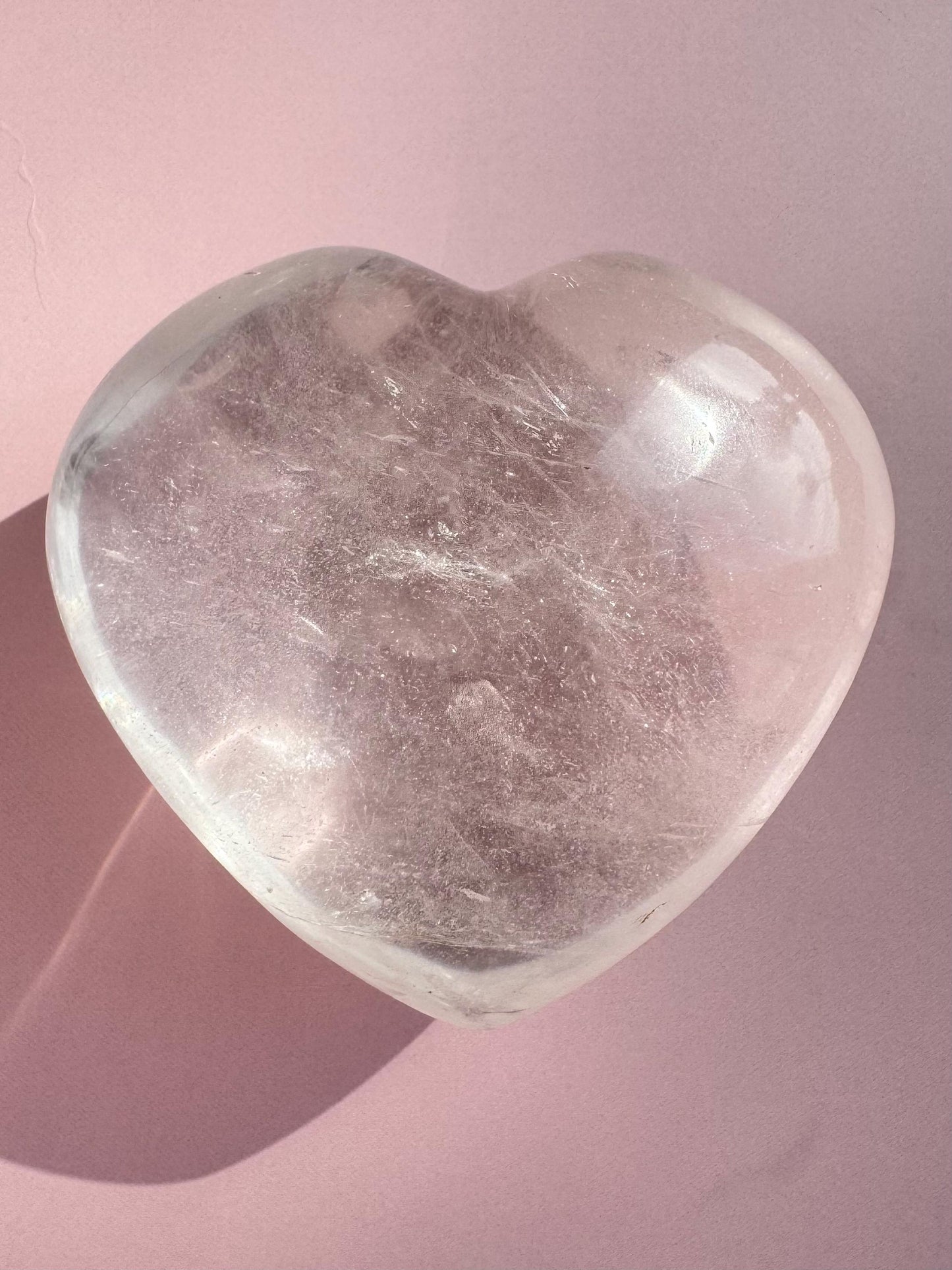 A Clear Quartz Heart Carving - Very High Clarity Crystal sits on a light pink surface, its heart-shaped form casting a gentle shadow to the left.