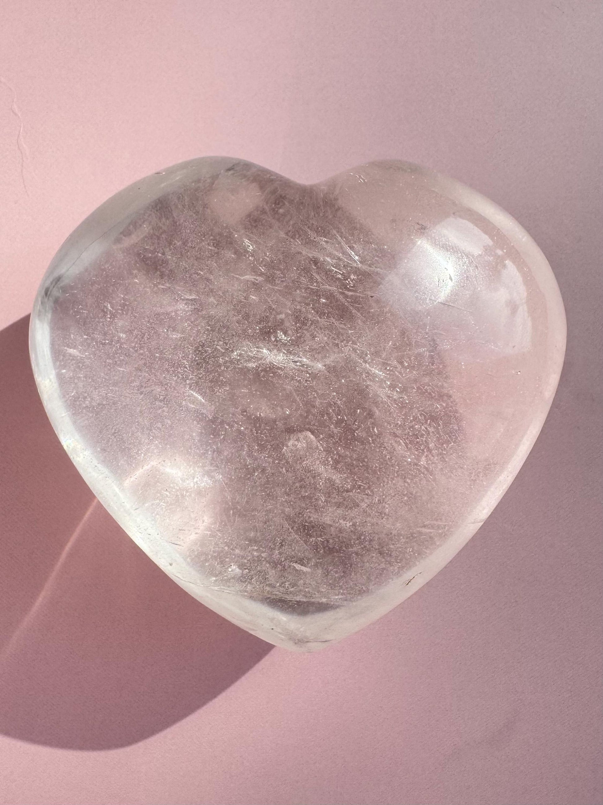 A Clear Quartz Heart Carving - Very High Clarity Crystal sits on a light pink surface, its heart-shaped form casting a gentle shadow to the left.