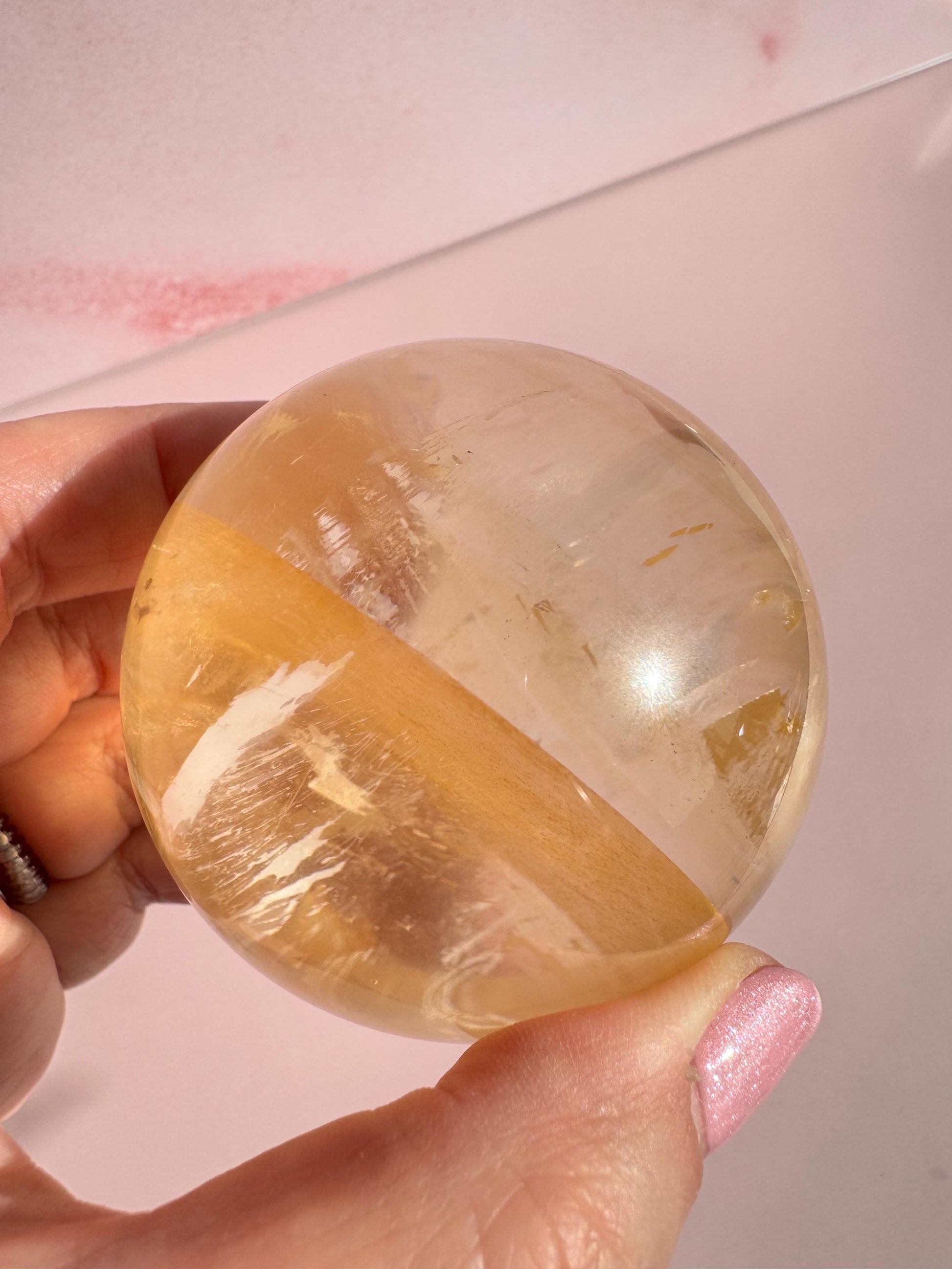 A hand holds the Honey Calcite Sphere (58 mm diameter), its yellow crystal bands gleaming with energizing beauty—perfect for motivation—set against a pale pink background.
