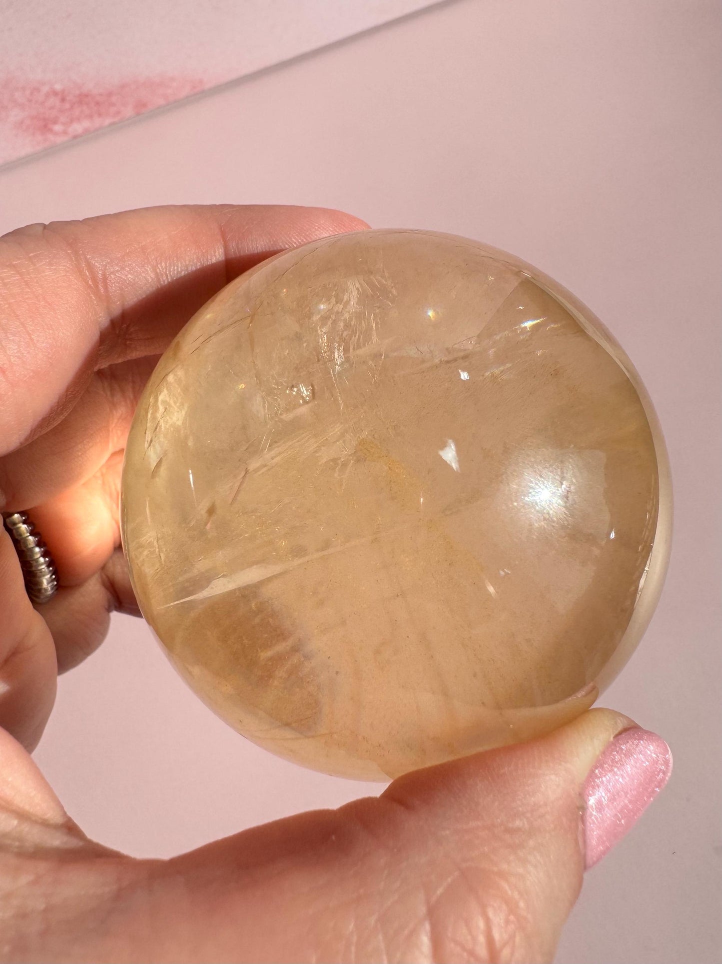 A hand holds the Honey Calcite Sphere (58 mm diameter), a crystal for motivation, against a pale pink background.