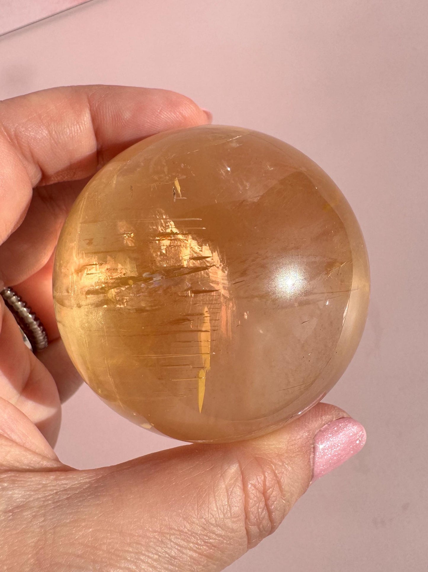 A hand holding a 58 mm Honey Calcite Sphere with visible internal striations, set against a light pink background—a captivating crystal for motivation and positive energy.