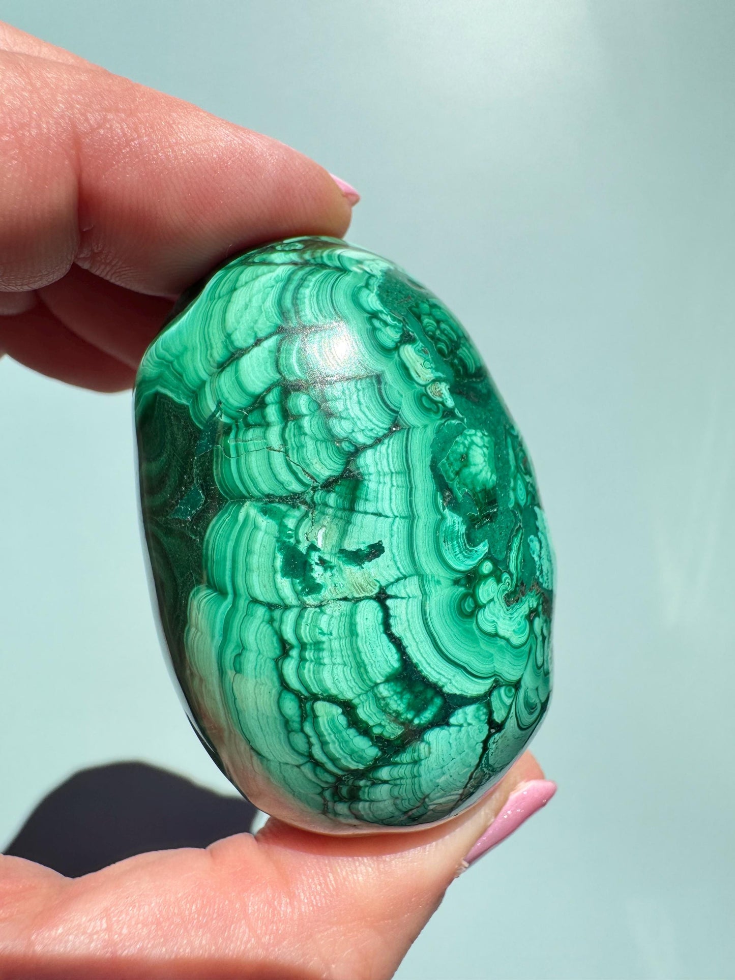 A hand holds the Malachite Egg-Shaped Carving (ITEM C), prized among healing crystals as a transformation stone, displaying green banded patterns against a light background.