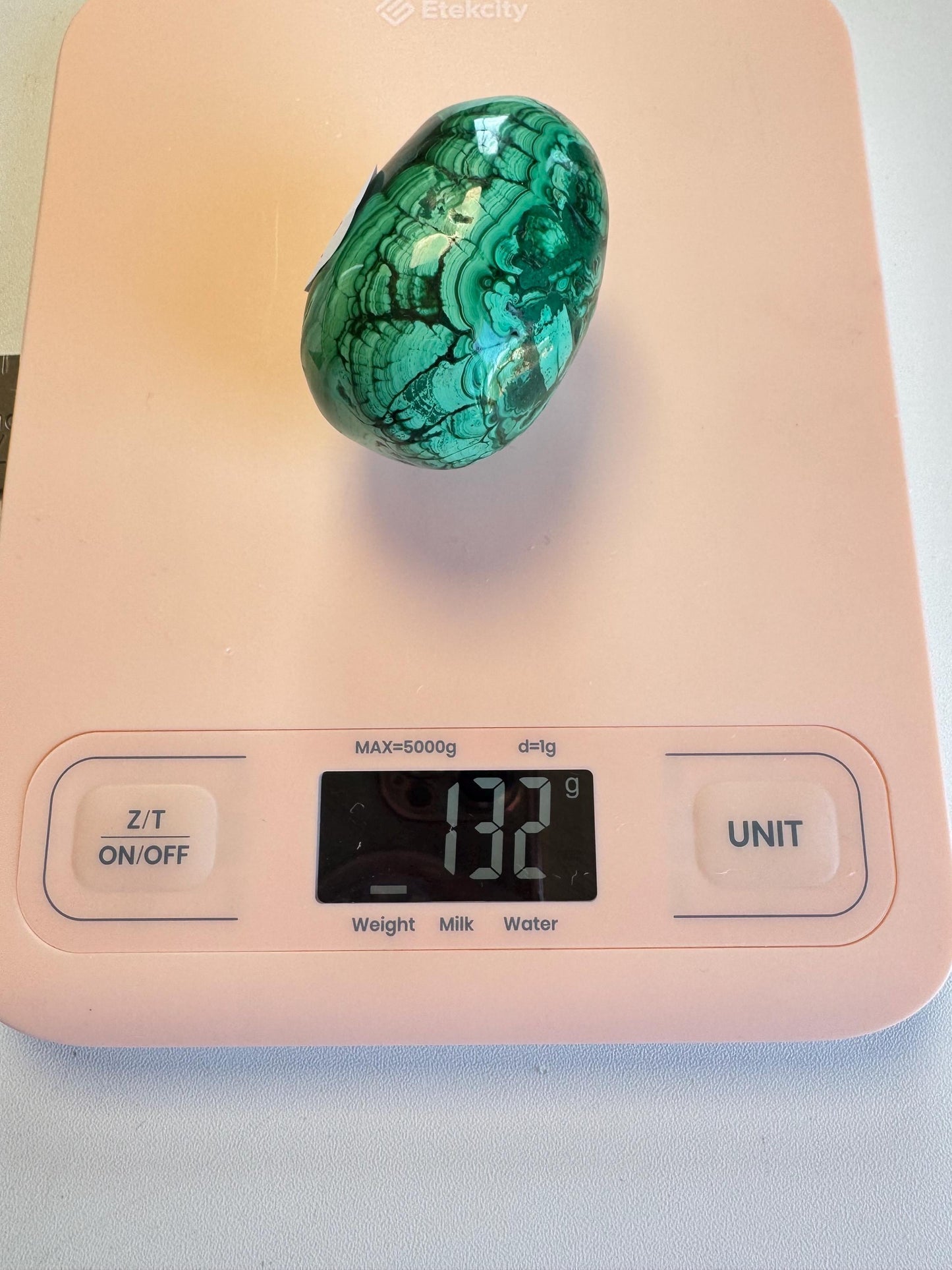 A Malachite Egg-Shaped Carving (ITEM C), prized among healing crystals as a transformation stone, sits on a pink digital kitchen scale showing a weight of 132 grams.