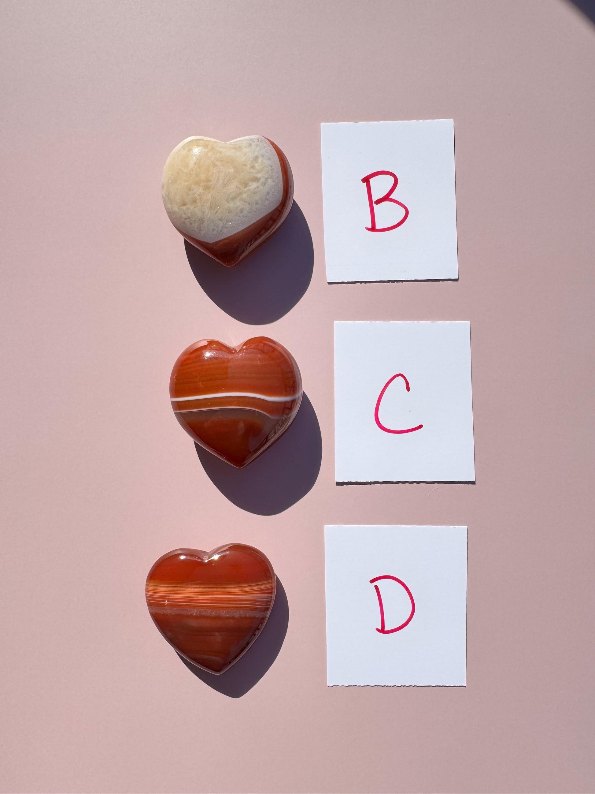 A Small Carnelian Heart Carving - YOU CHOOSE is arranged vertically with two other heart-shaped stones on a pink surface beside cards labeled B, C, and D in red. These crystals bring vibrant energy to the display.