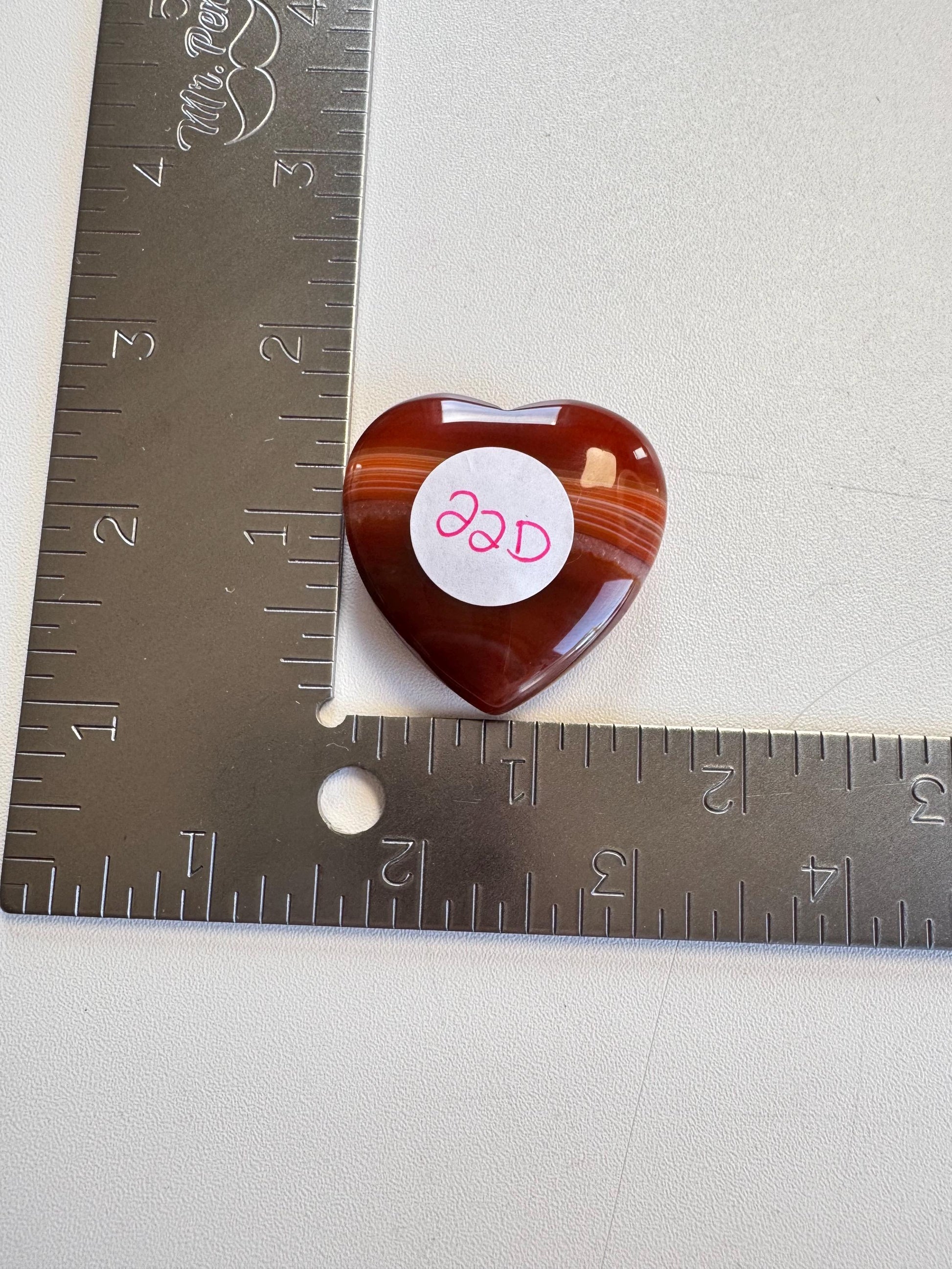 A Small Carnelian Heart Carving - YOU CHOOSE rests at the corner of a metal ruler marked in inches, making it a unique energizing stone for collectors and enthusiasts.