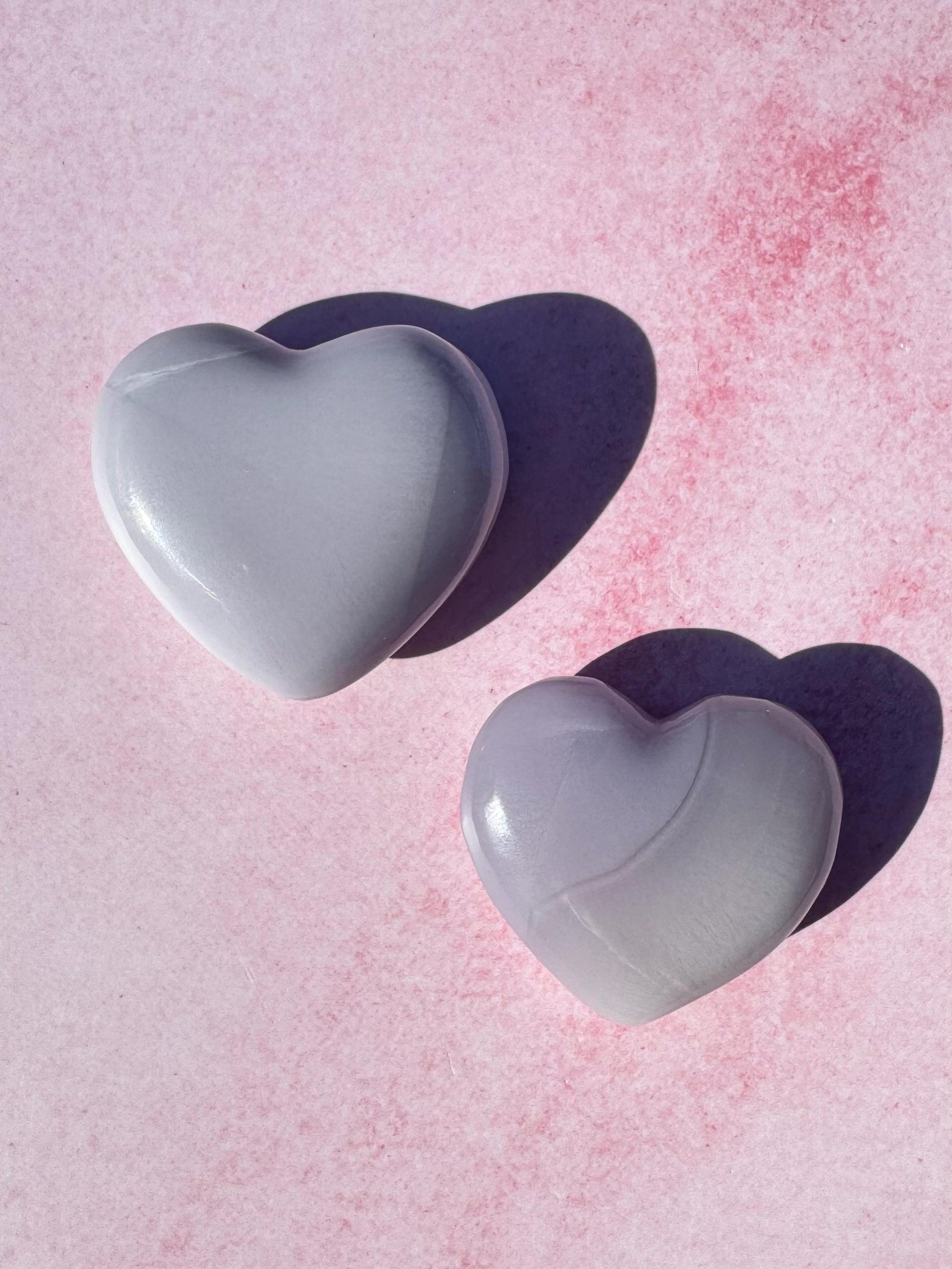 Two Lavender Fluorite Heart Carvings from Mexico—YOU CHOOSE—are placed on a pink textured surface, casting soft leftward shadows.