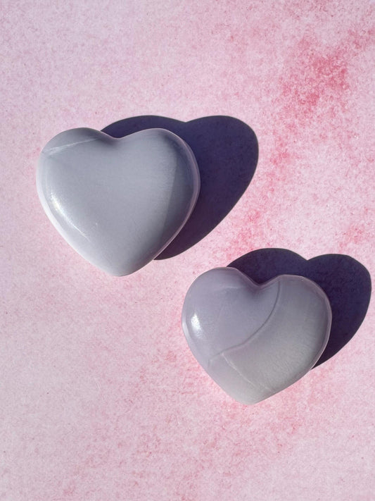 Two Lavender Fluorite Heart Carvings from Mexico—YOU CHOOSE—are placed on a pink textured surface, casting soft leftward shadows.